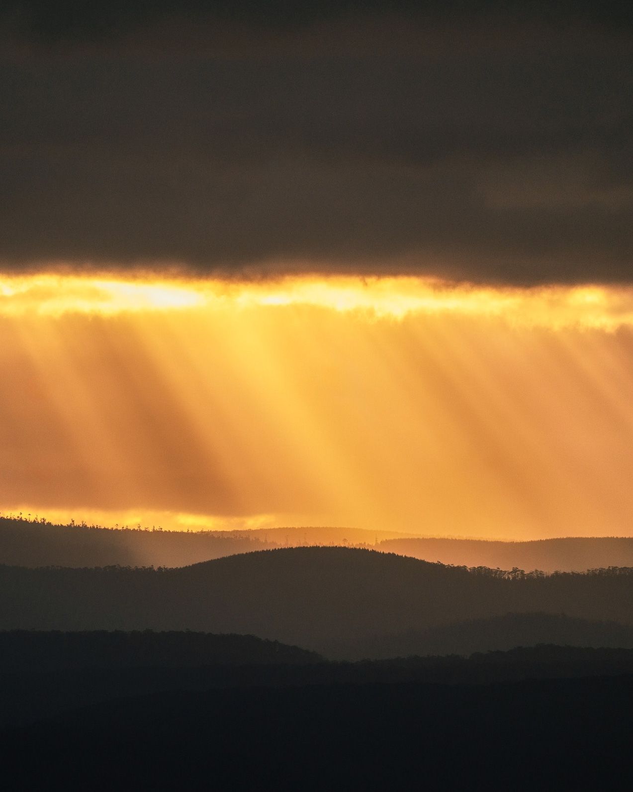 Golden sunbeams pierce dramatic storm clouds over silhouetted rolling hills at sunset.