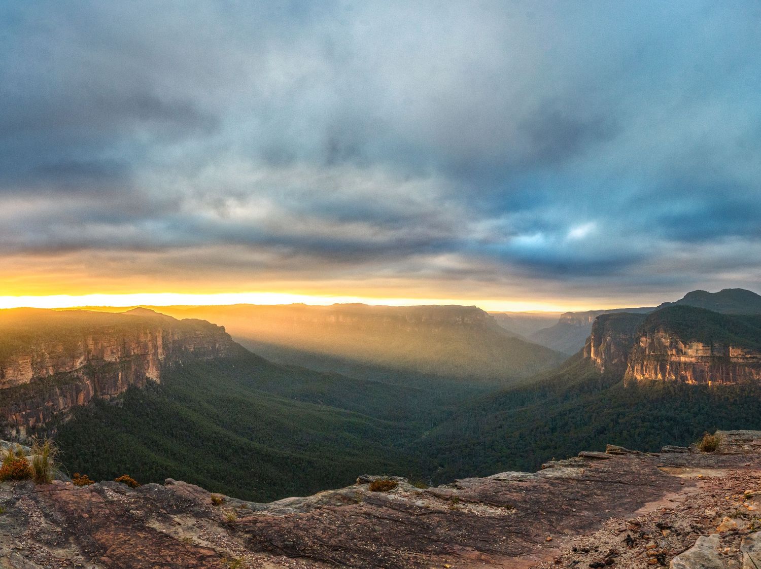 Panoramic sunset view over Blue Mountains valley with dramatic clouds, golden light, and lush green canyon landscape.