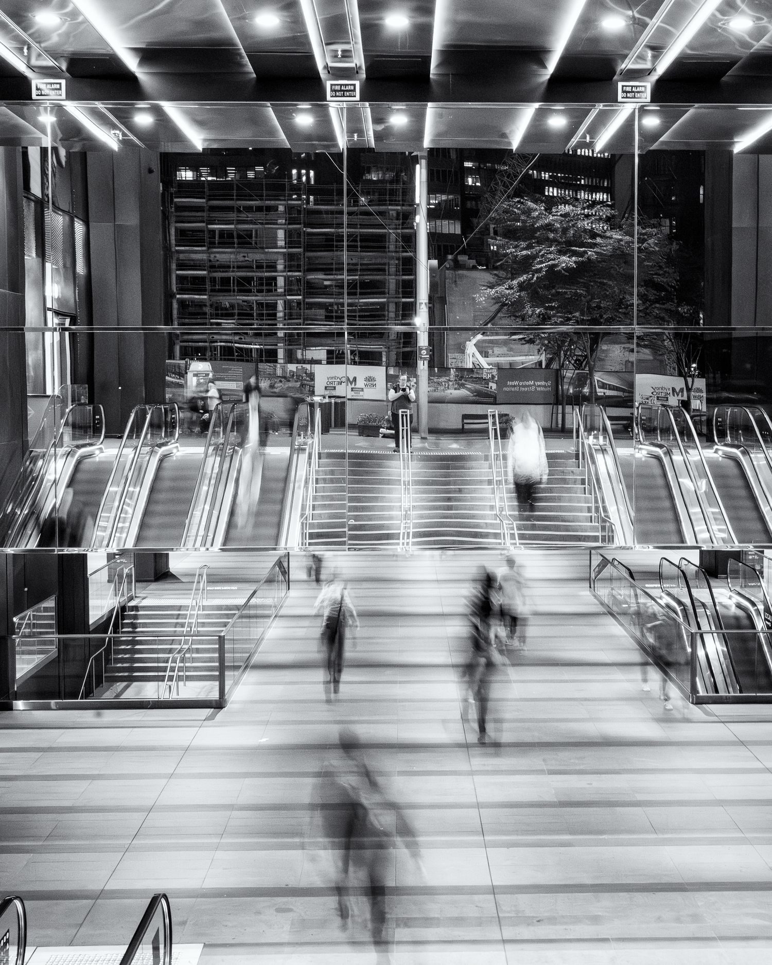 Black and white long exposure photo of blurred commuters on escalators and stairs in a modern transit hub at night.