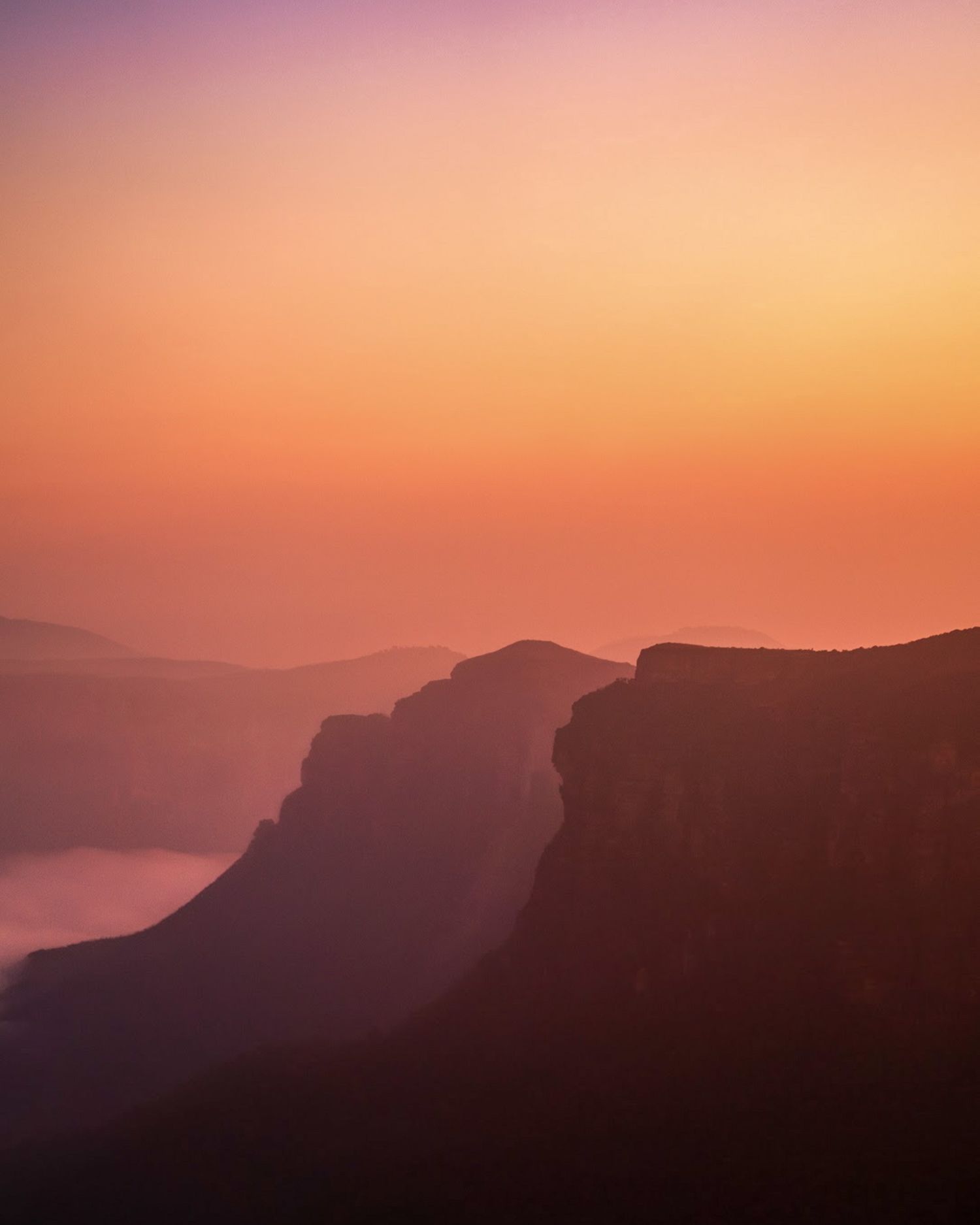 Stunning sunset over misty mountain canyon with vibrant orange and purple gradient sky casting silhouettes on rugged cliffs.