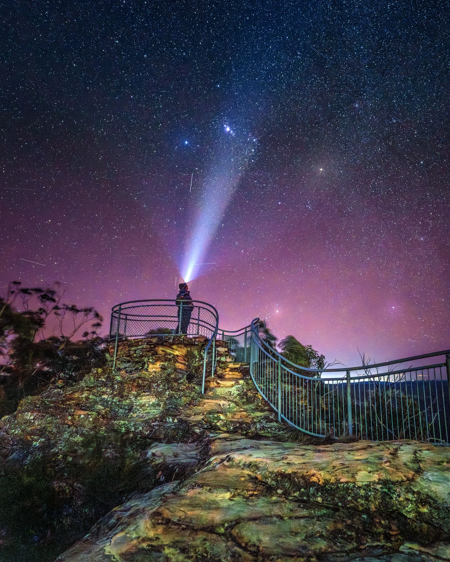 Person standing on rocky lookout shining headlamp at starry night sky with purple hues and Milky Way visible.