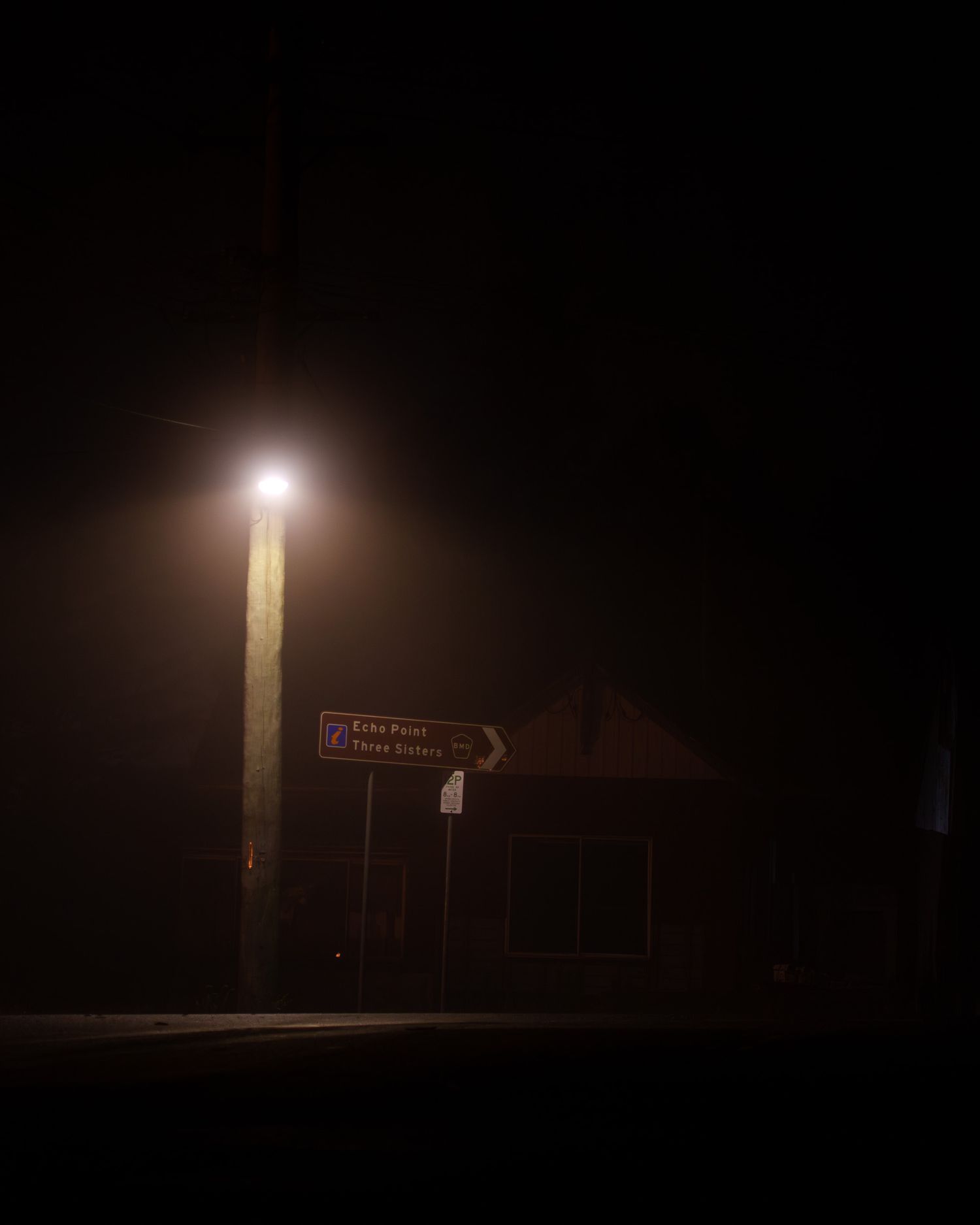 A dimly lit street corner at night with a glowing streetlight illuminating a directional road sign on a wooden pole.