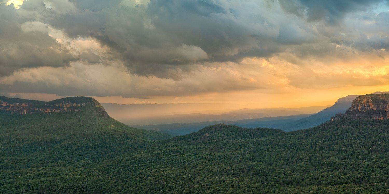 Panoramic sunset view over lush green mountains with dramatic storm clouds and golden light rays in Blue Mountains, Australia.