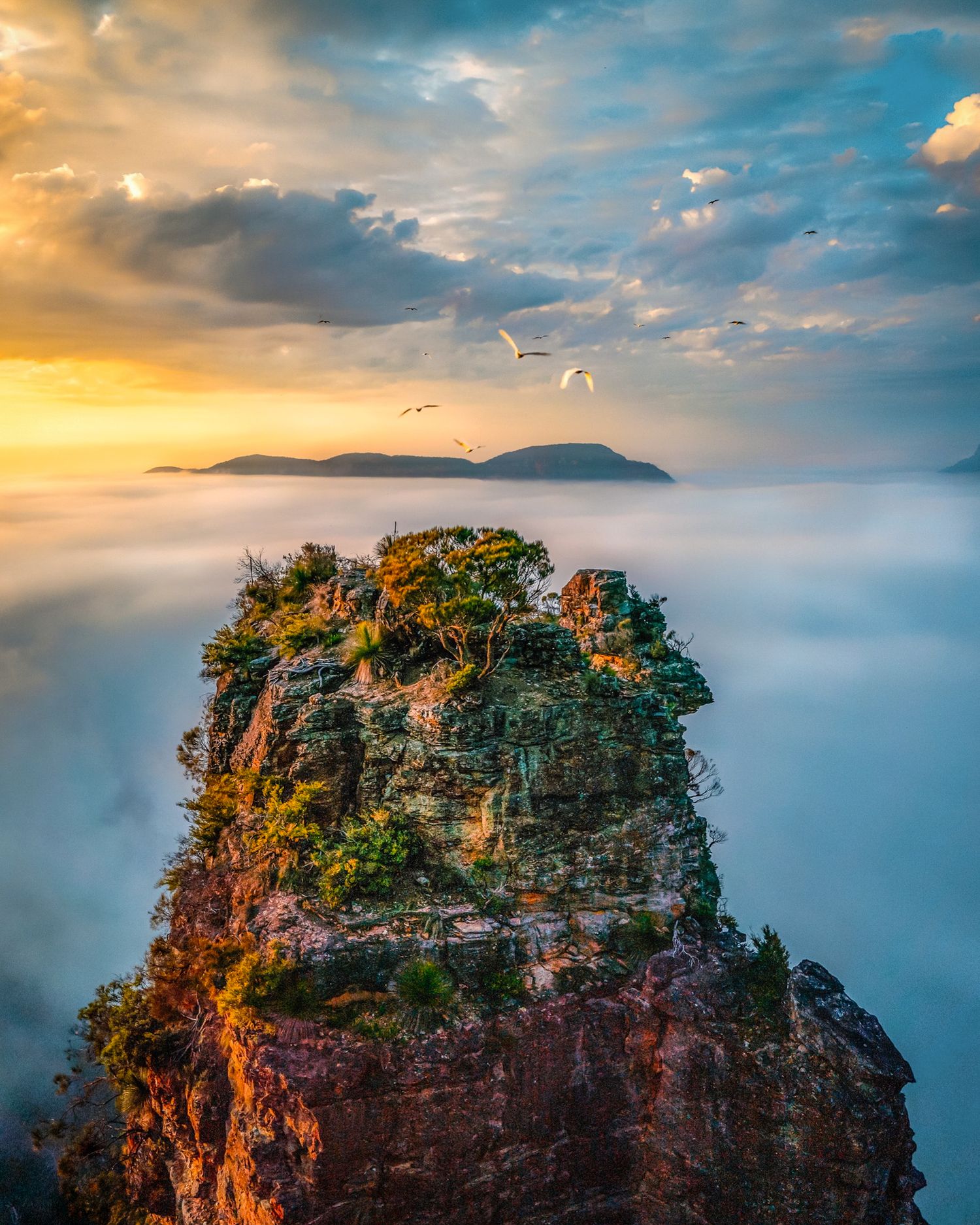 Rock pinnacle emerges from sea of clouds during dramatic sunset with birds soaring overhead.