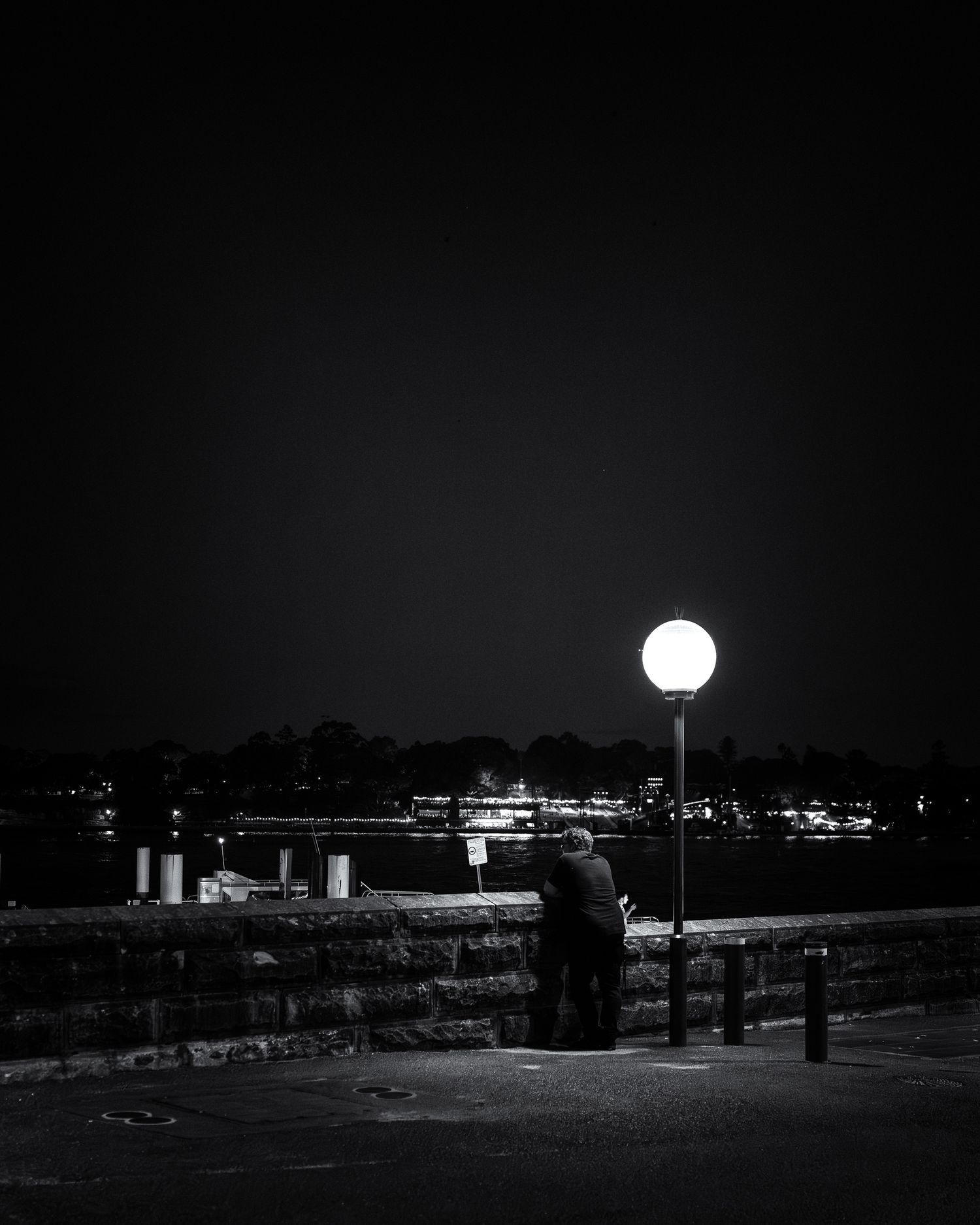 A lone person stands under a glowing street lamp at night by a waterfront in this moody black and white photo.