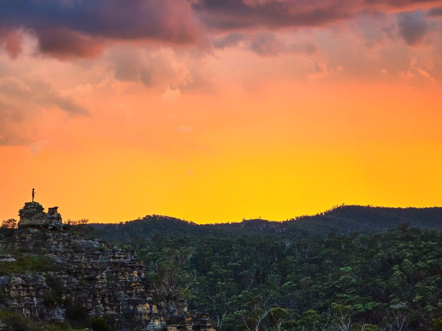 Vibrant orange sunset sky over silhouetted rocky cliffs and lush green forest hills in a dramatic landscape.