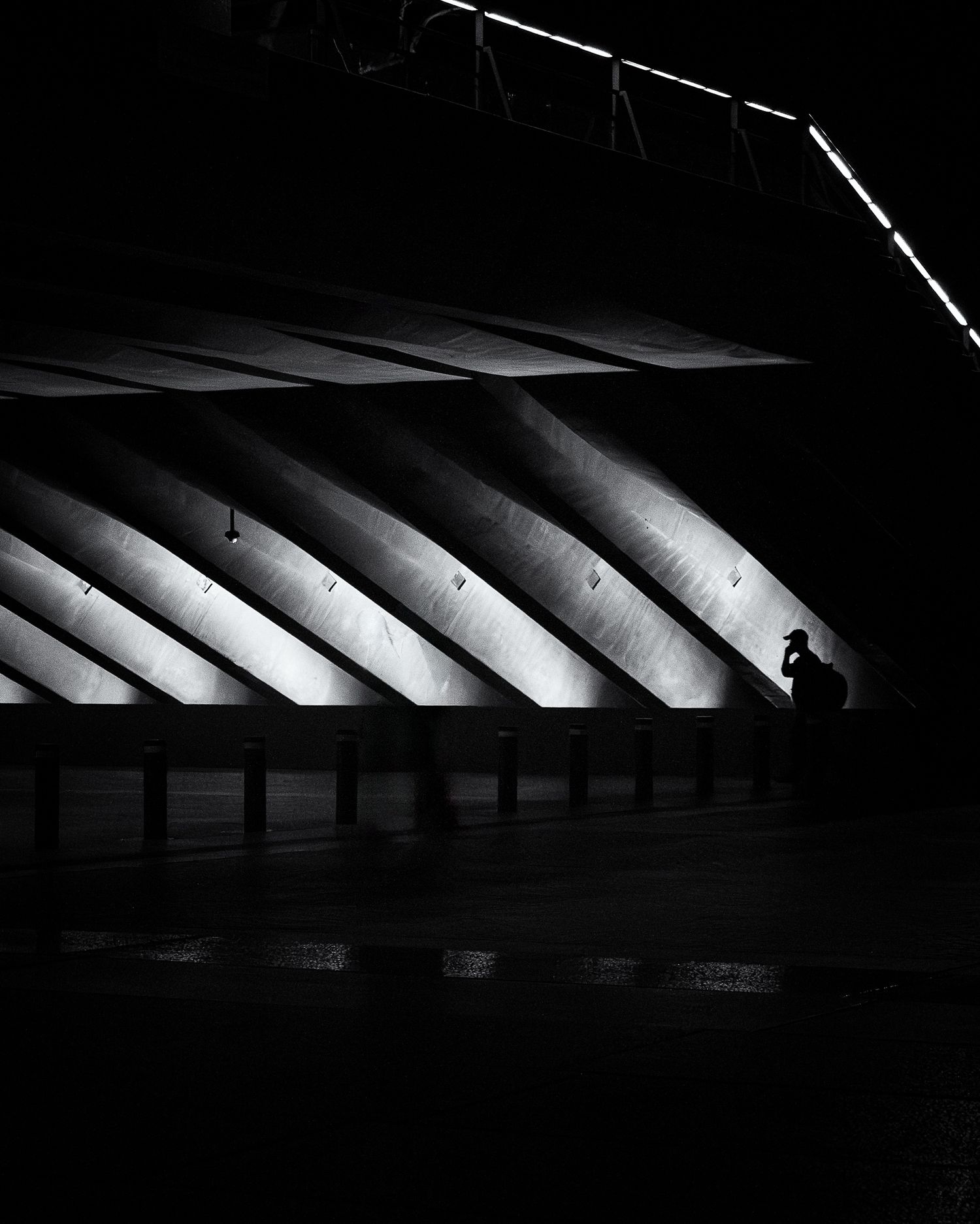 Black and white photo of a lone silhouette sitting beneath dramatic diagonal concrete arches in dark urban architecture.