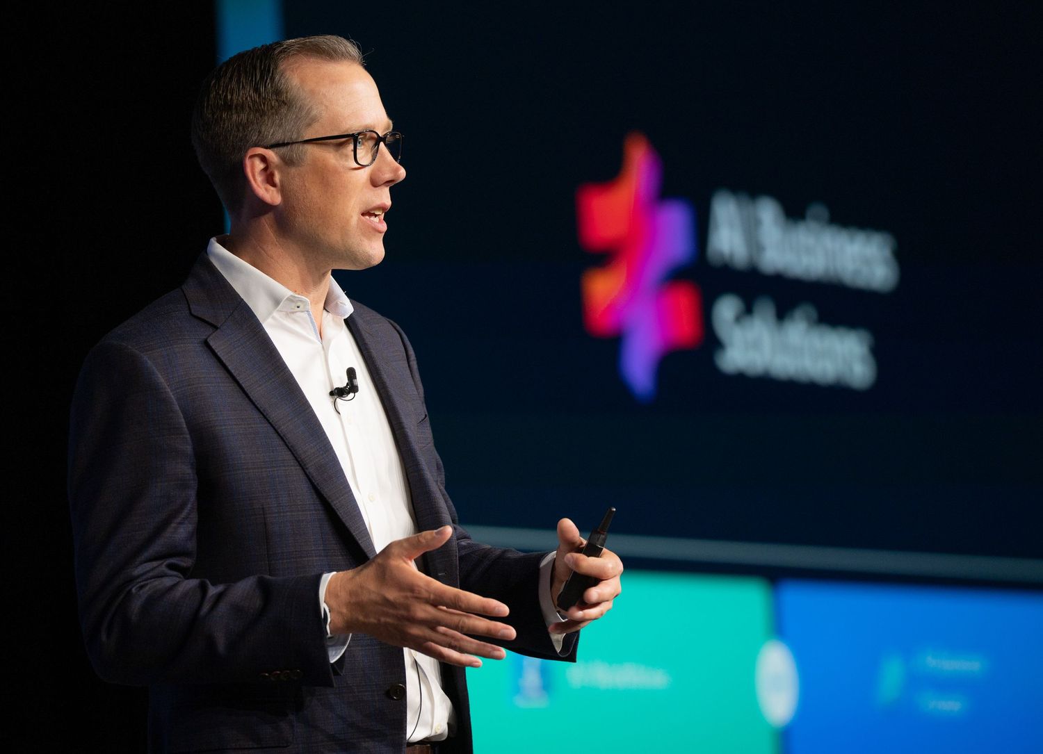 A speaker in a dark suit gestures while presenting on stage with a blue and teal backdrop.