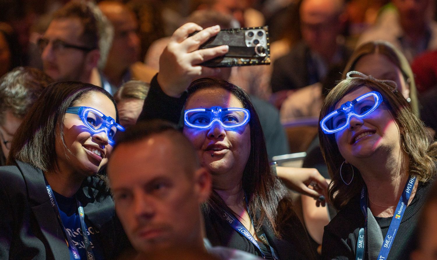 Audience members wearing glowing blue LED glasses smile and enjoy an event in a dark venue.