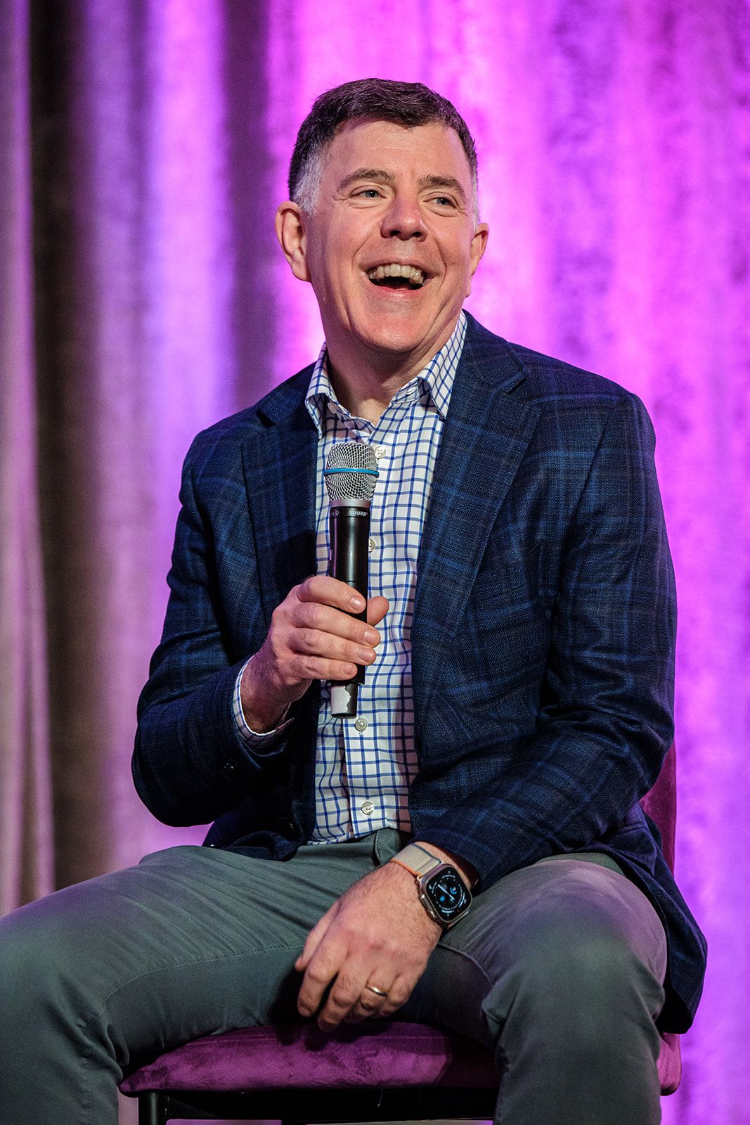 A speaker in a blue blazer and checkered shirt sits on stage with purple lighting while holding a microphone.