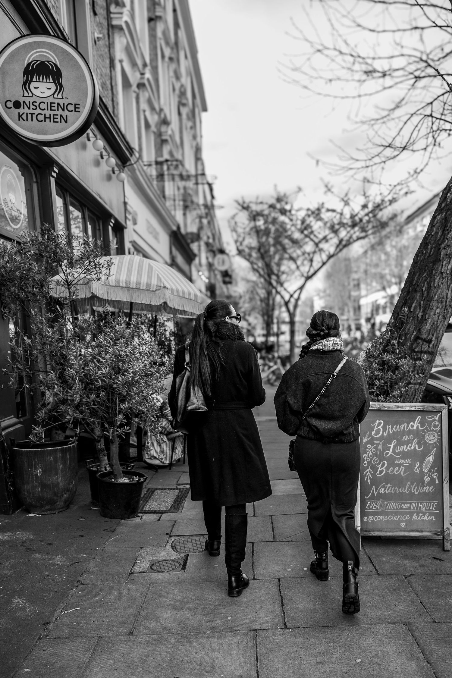 Two people walk past storefronts and chalkboard signs on a city sidewalk in black and white photography.