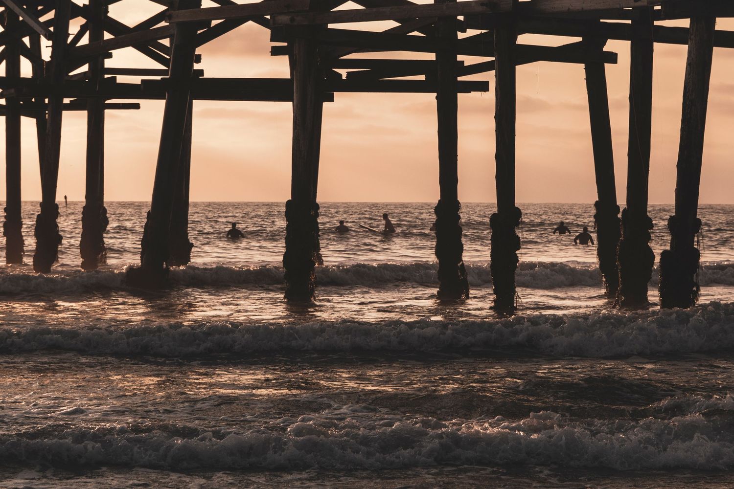 Silhouetted wooden pier pilings stand against a moody sunset sky reflected in calm ocean waters.