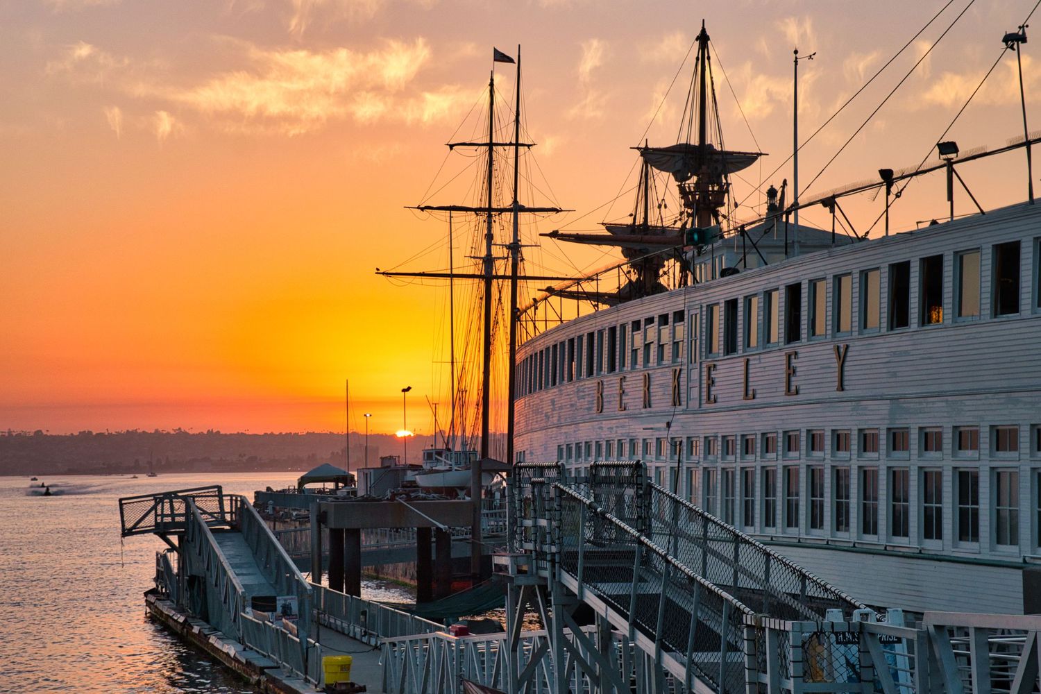 Boats and yachts docked at a marina during a vibrant orange sunset with silhouetted masts against the sky.