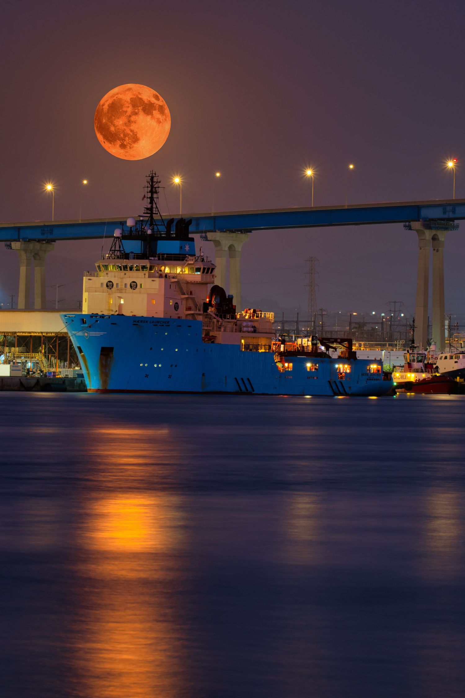 Naval ships docked at harbor under full orange moon with city lights reflecting on water.