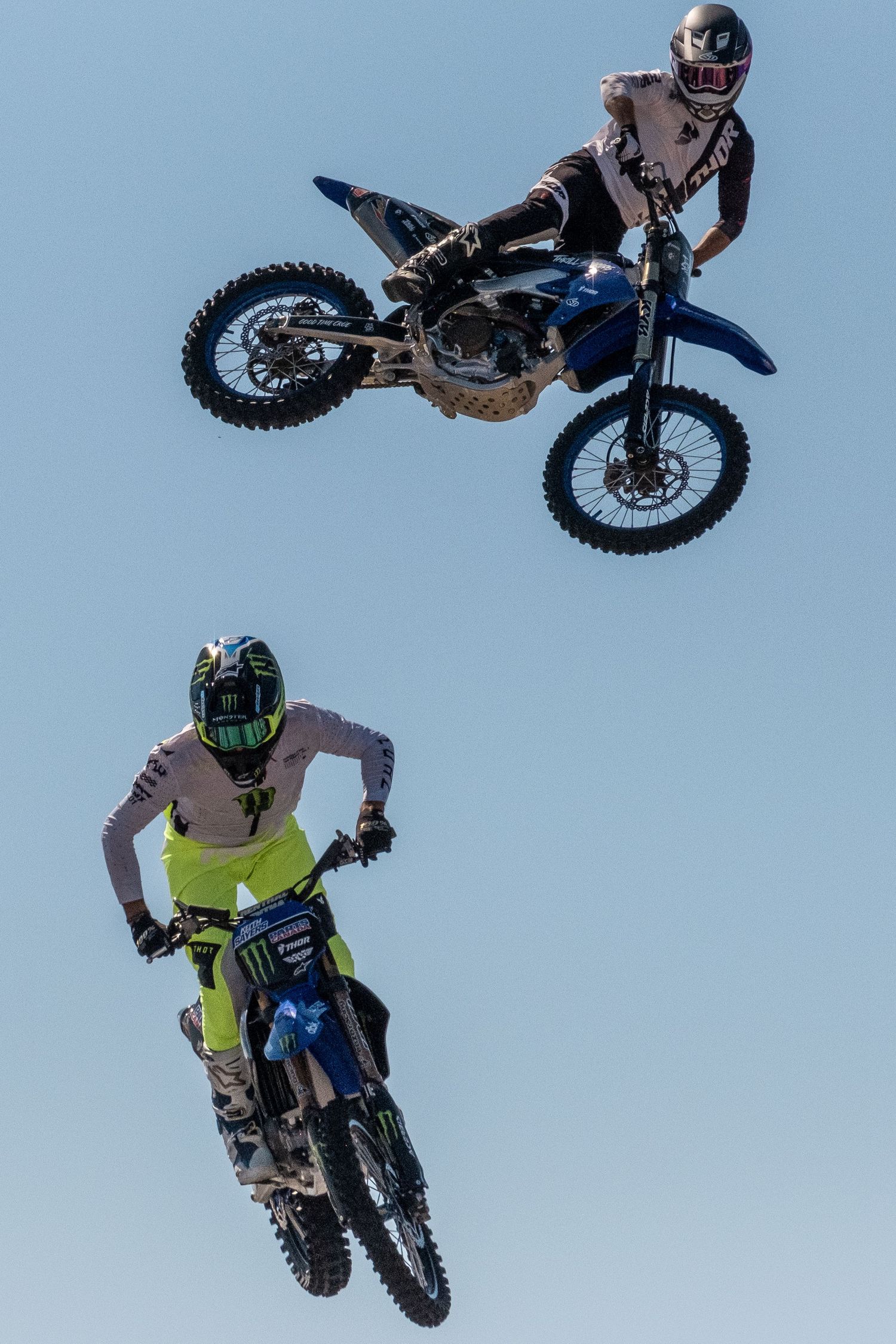 Motocross riders performing aerial stunts and jumps against clear blue sky.