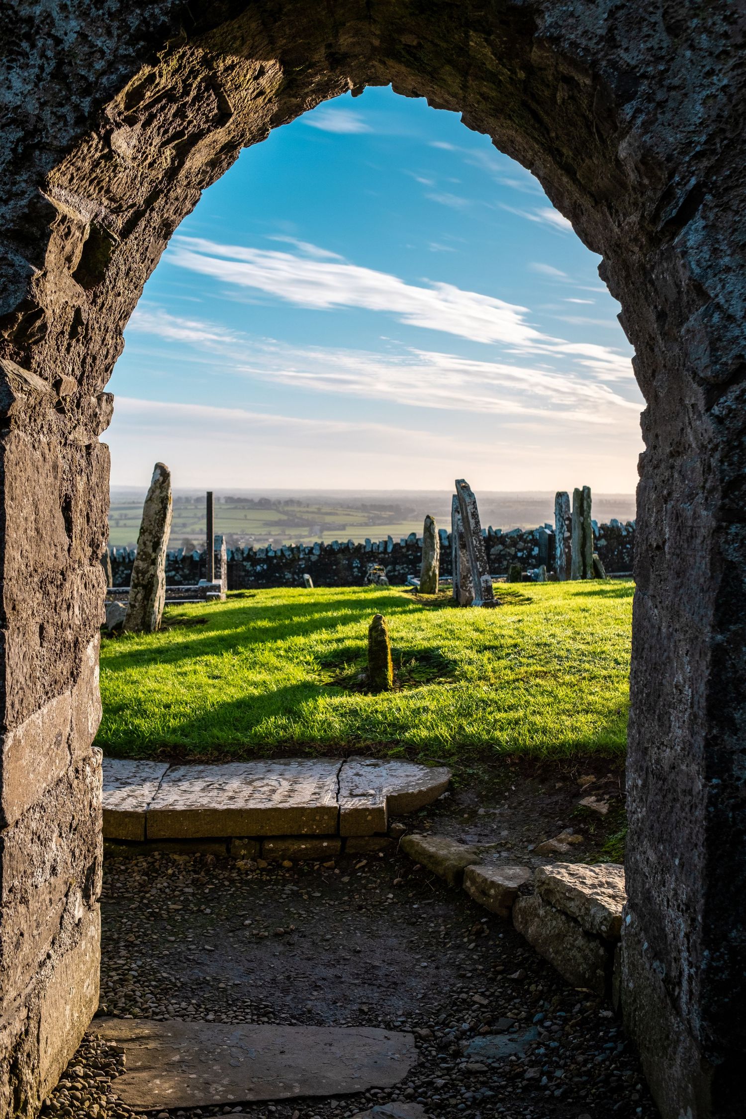Stone archway frames a scenic view of ancient standing stones on a grassy hilltop against a bright blue sky.