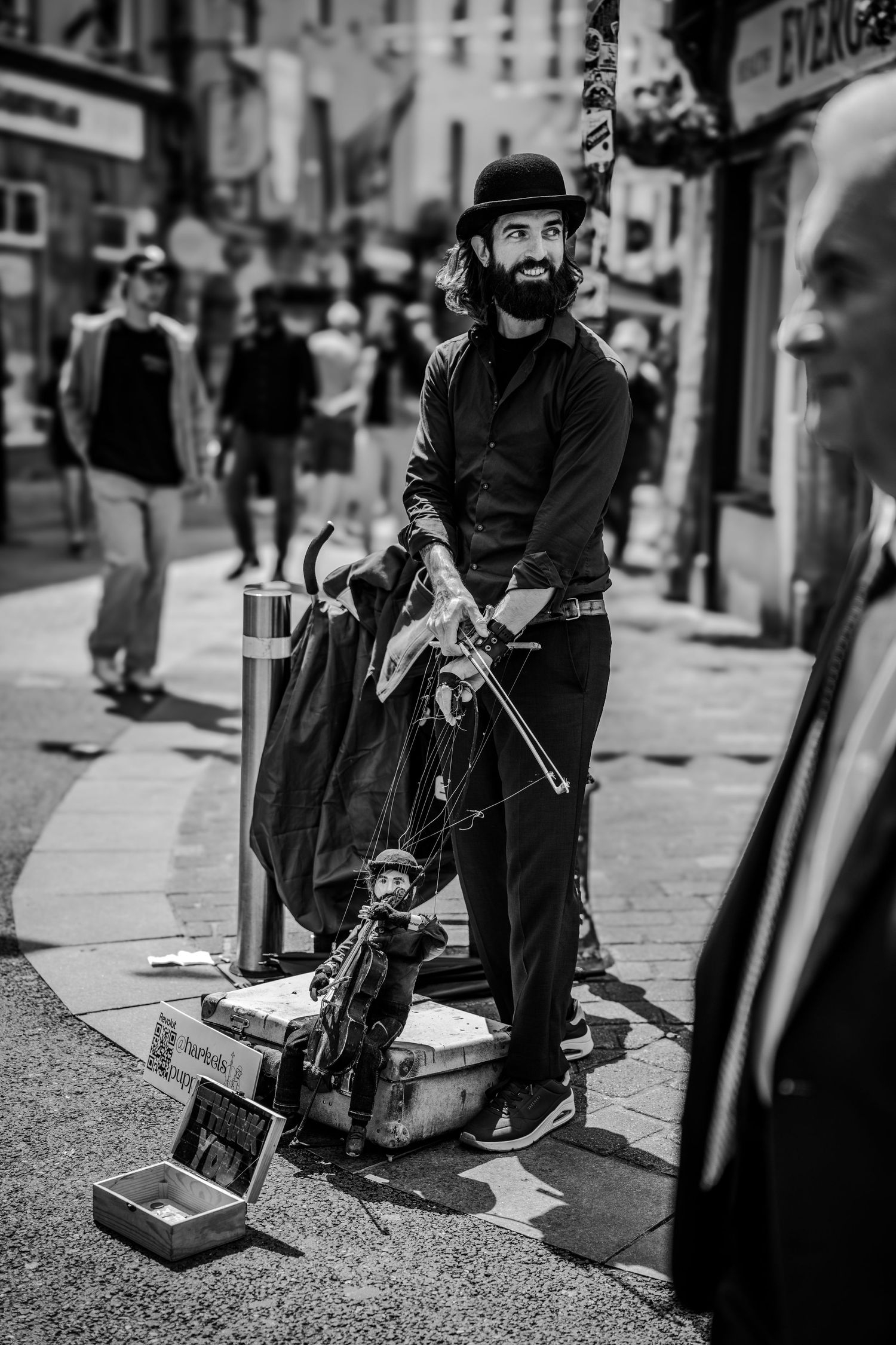 A black and white street photograph of a musician performing with a guitar case open for tips on a city sidewalk.