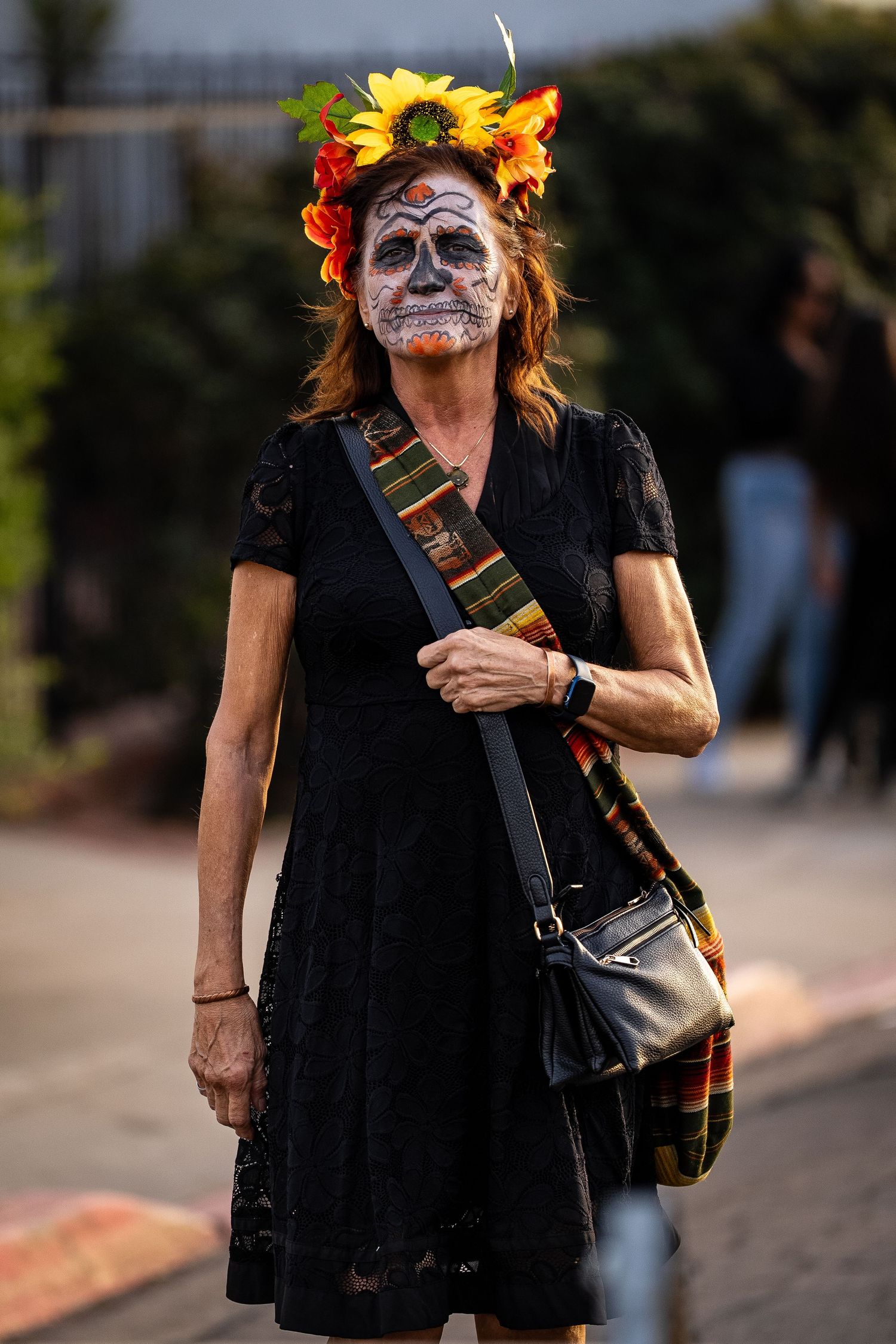Person in black dress with Day of the Dead makeup and orange floral headpiece walking outdoors.
