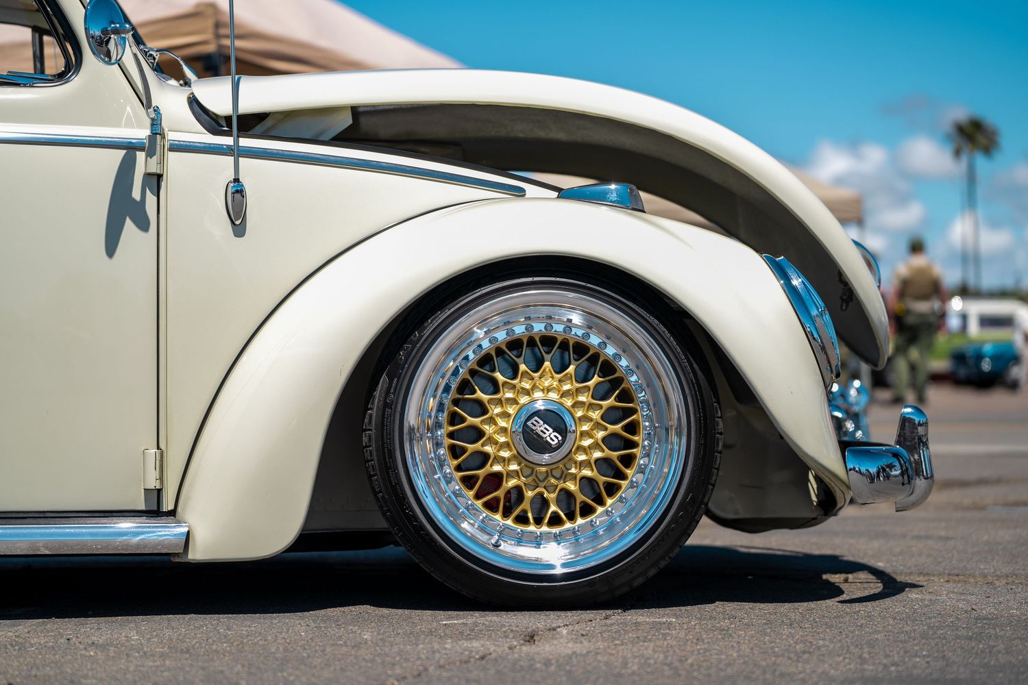 Close-up of a classic cream VW Beetle featuring a gold and chrome BBS wheel at a car show.