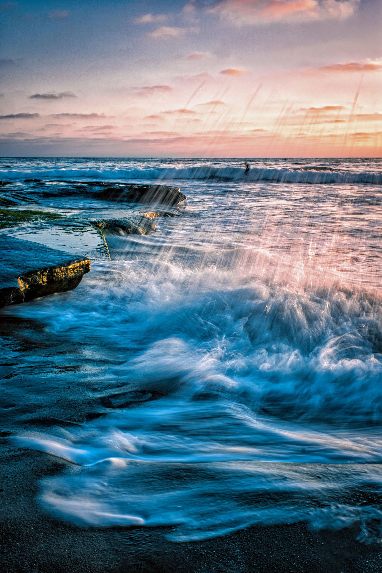Dramatic ocean waves crashing over rocky coastline during pink and blue sunset.