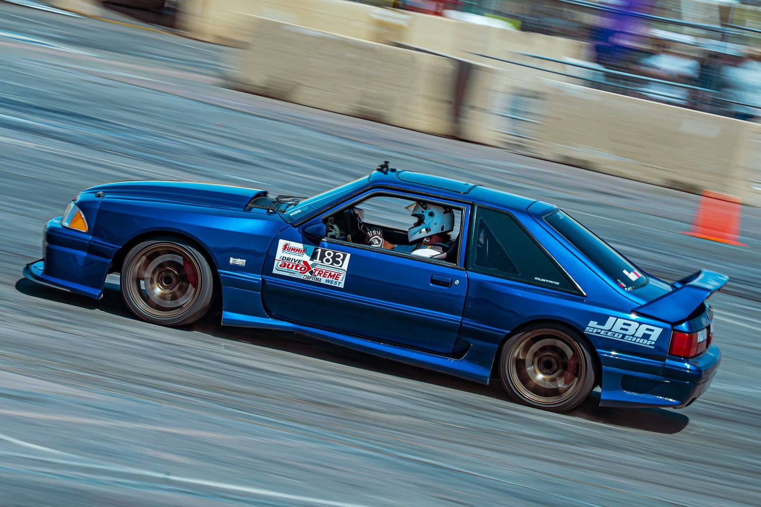 Blue Ford Mustang Fox Body race car drifting on a track during a motorsport competition event.