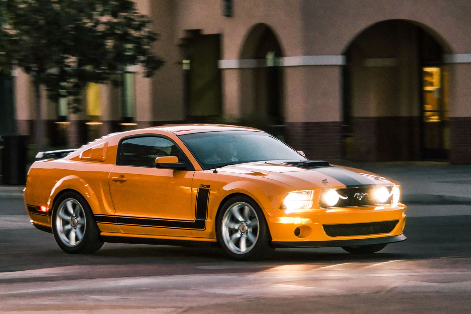 Orange Ford Mustang GT with racing stripes photographed in motion at dusk.