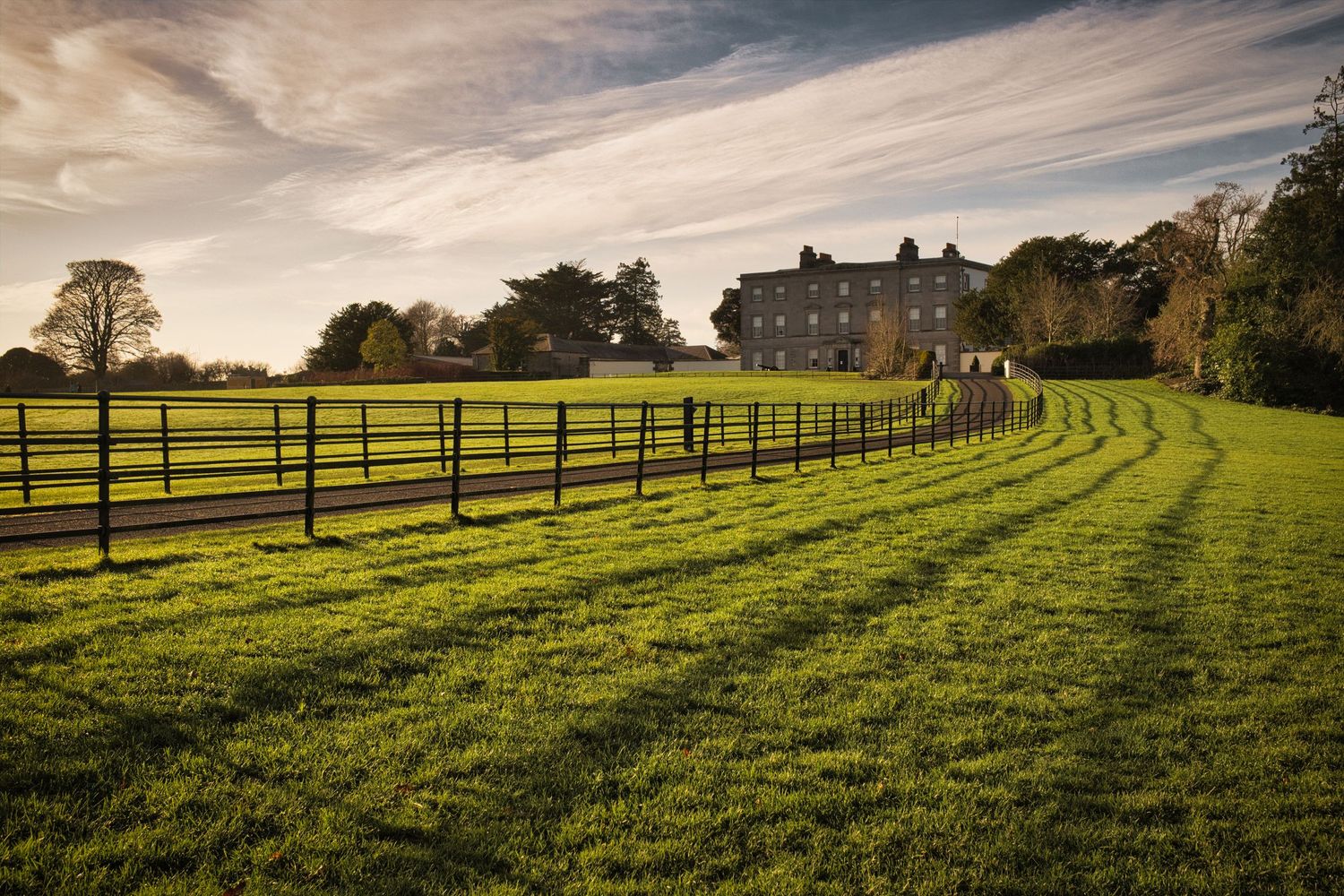 Elegant country estate with curved fence lines and green pastures at sunset casting long shadows across manicured grounds.