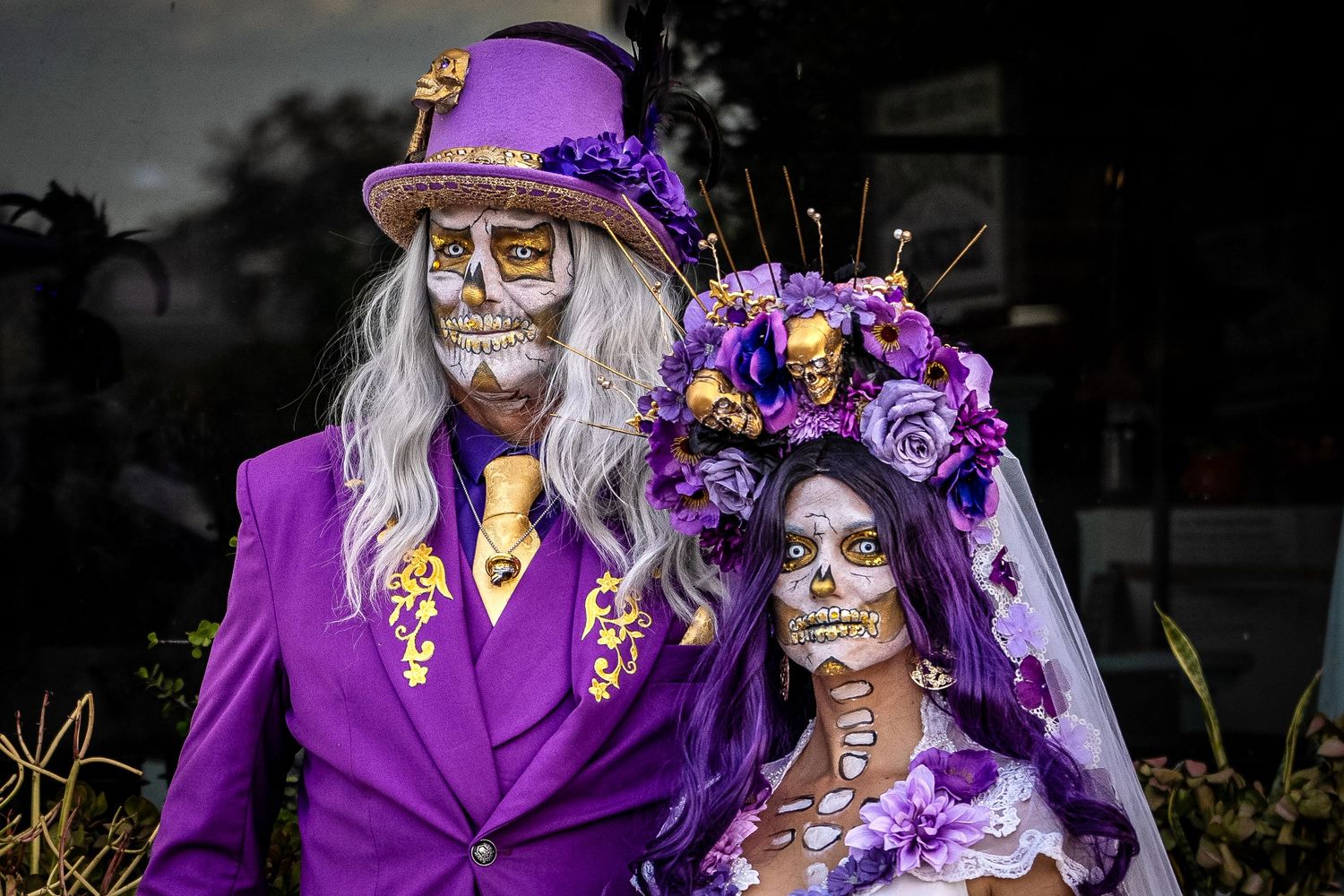 Day of the Dead themed couple in purple attire with sugar skull makeup and floral decorations.