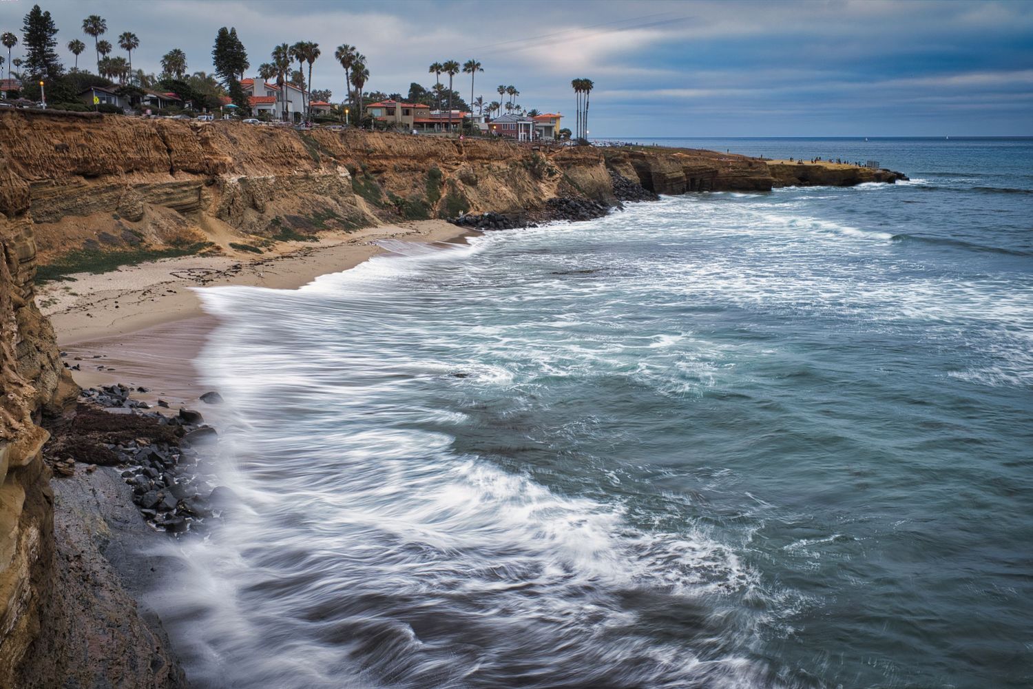 Panoramic coastal landscape with crashing waves against rocky cliffs under cloudy skies.
