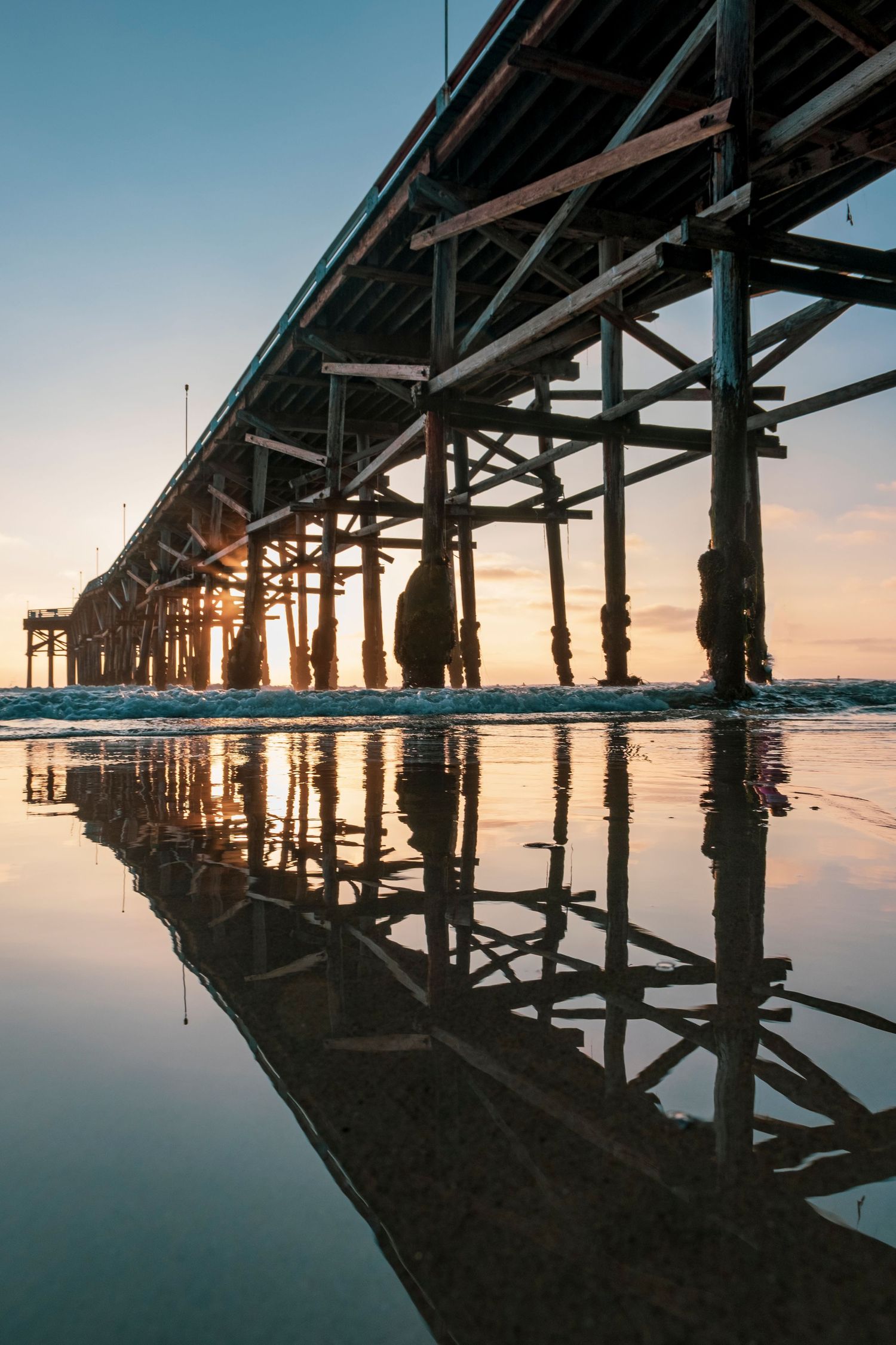 Wooden pier reflected in calm beach water during sunset creating perfect mirror image.