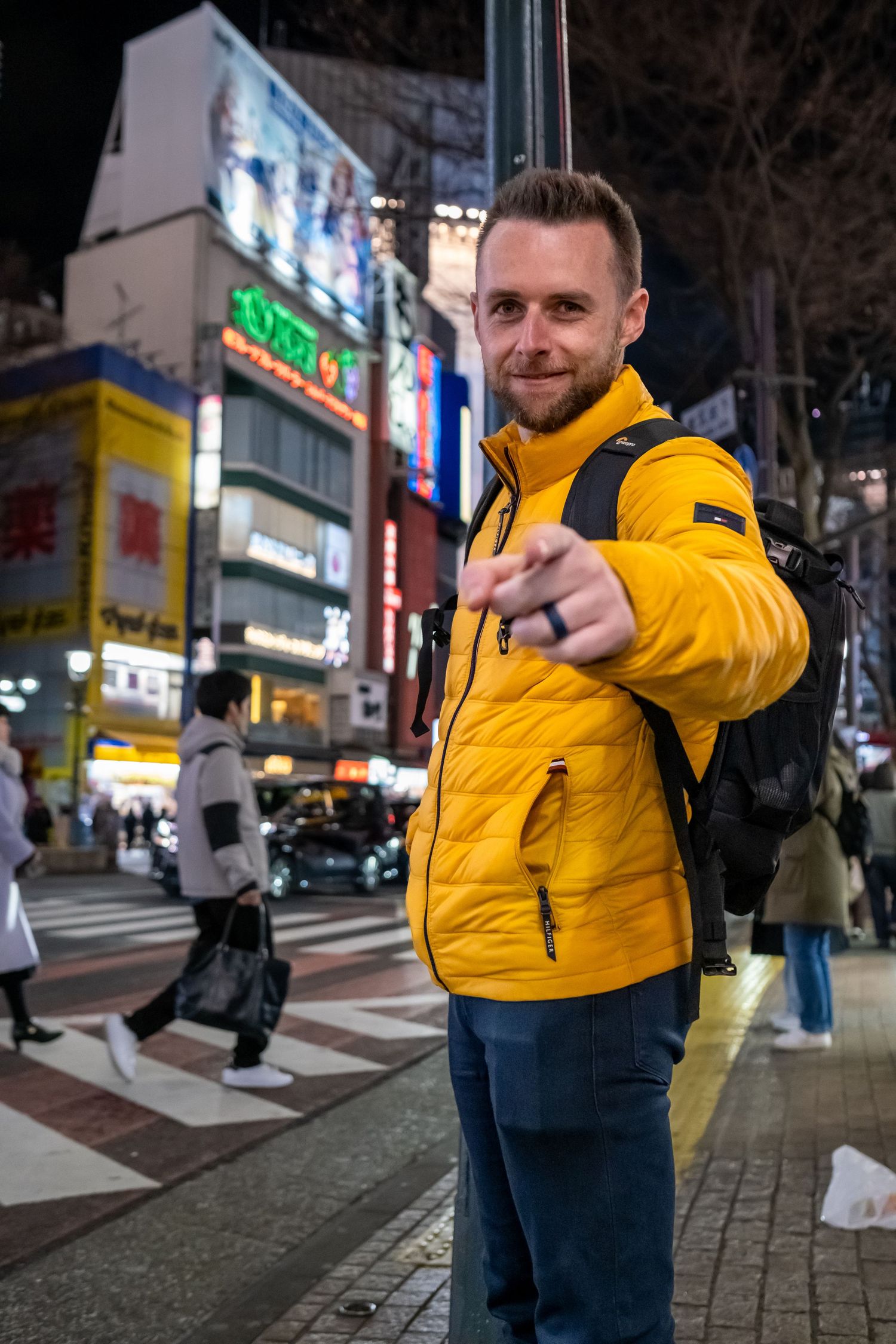 Person in bright yellow puffer jacket poses playfully on nighttime city street with illuminated storefronts in background.