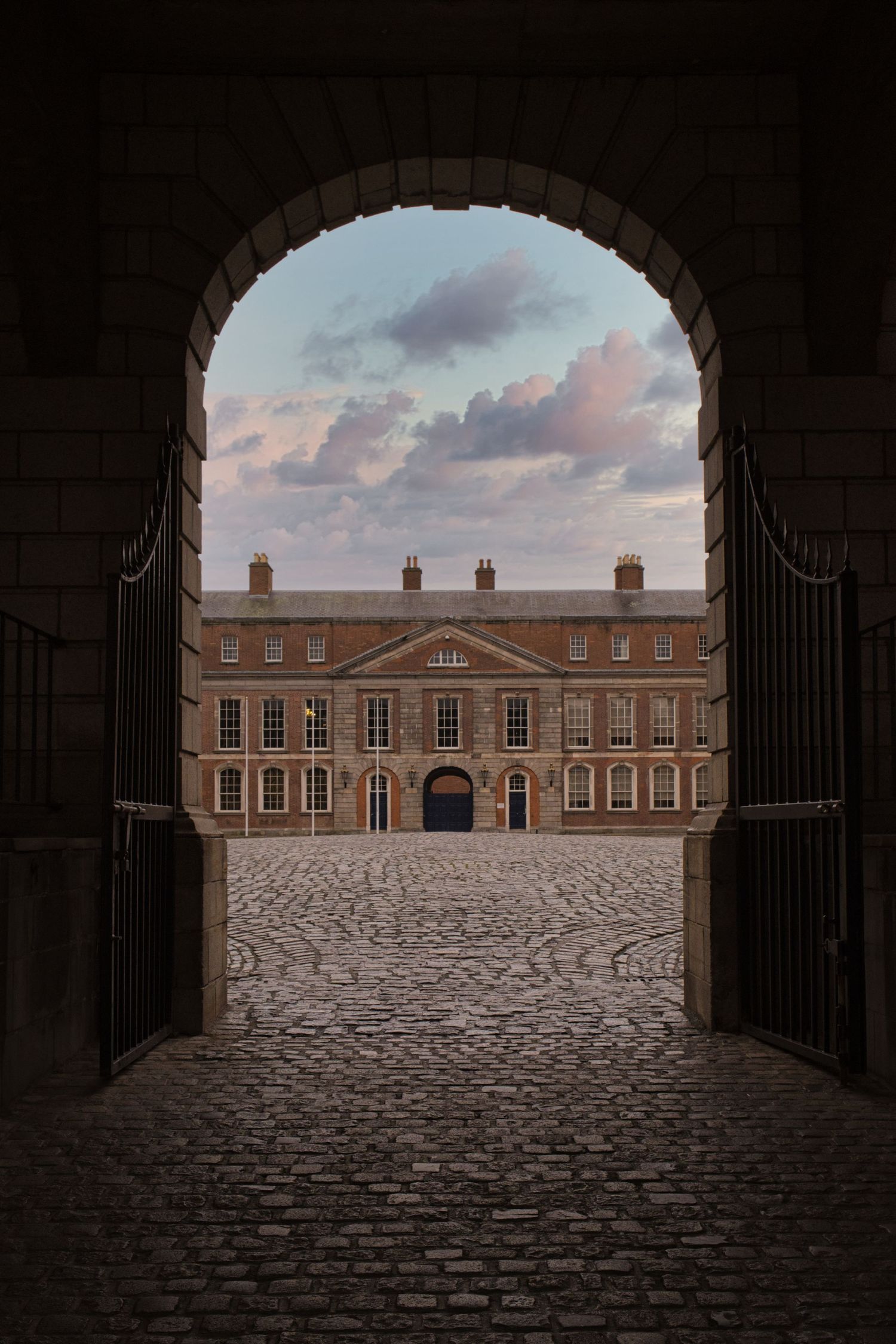 Historic brick building viewed through arched gateway with cobblestone courtyard.