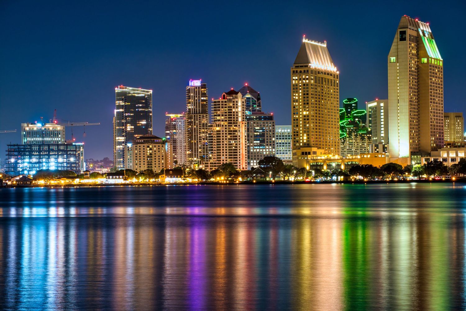 Panoramic nighttime cityscape showing illuminated skyscrapers reflecting colorfully on calm waterfront.