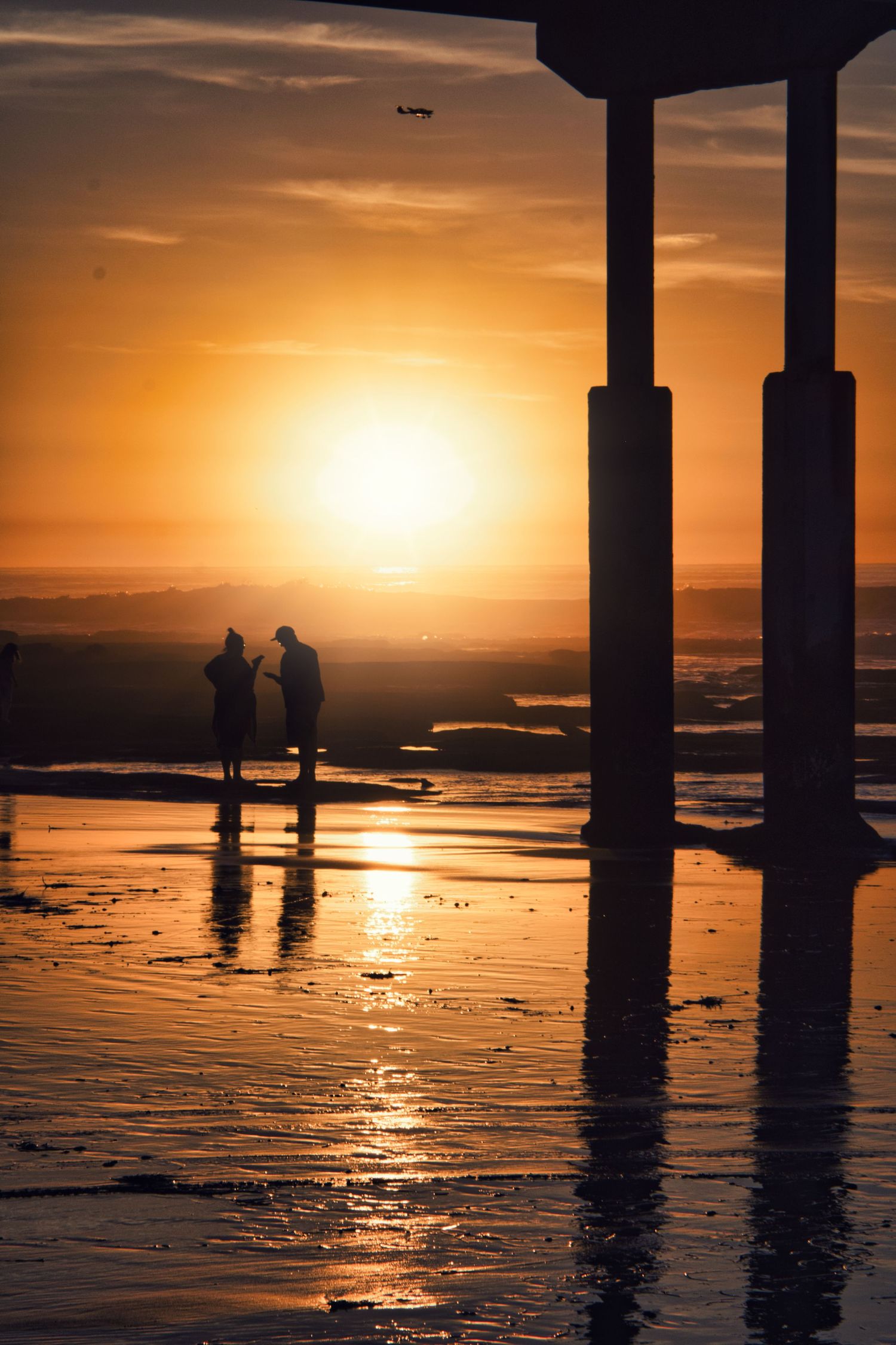 Dramatic beach sunset with silhouettes of pillars and reflections on wet sand.