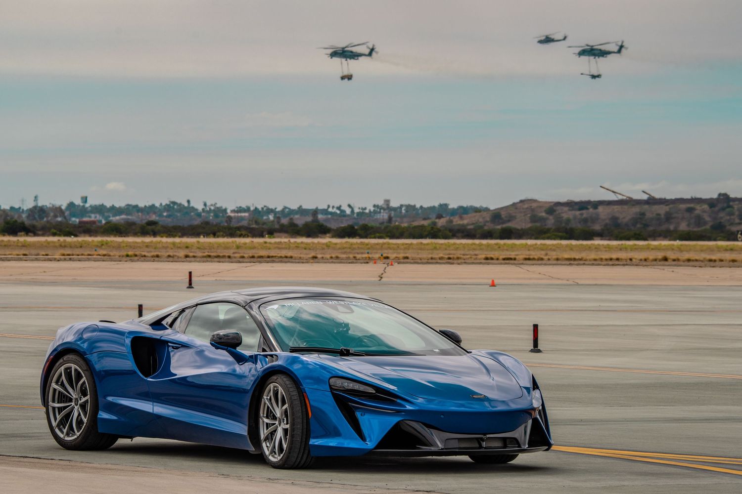 Blue McLaren supercar parked on airfield with helicopters in background.