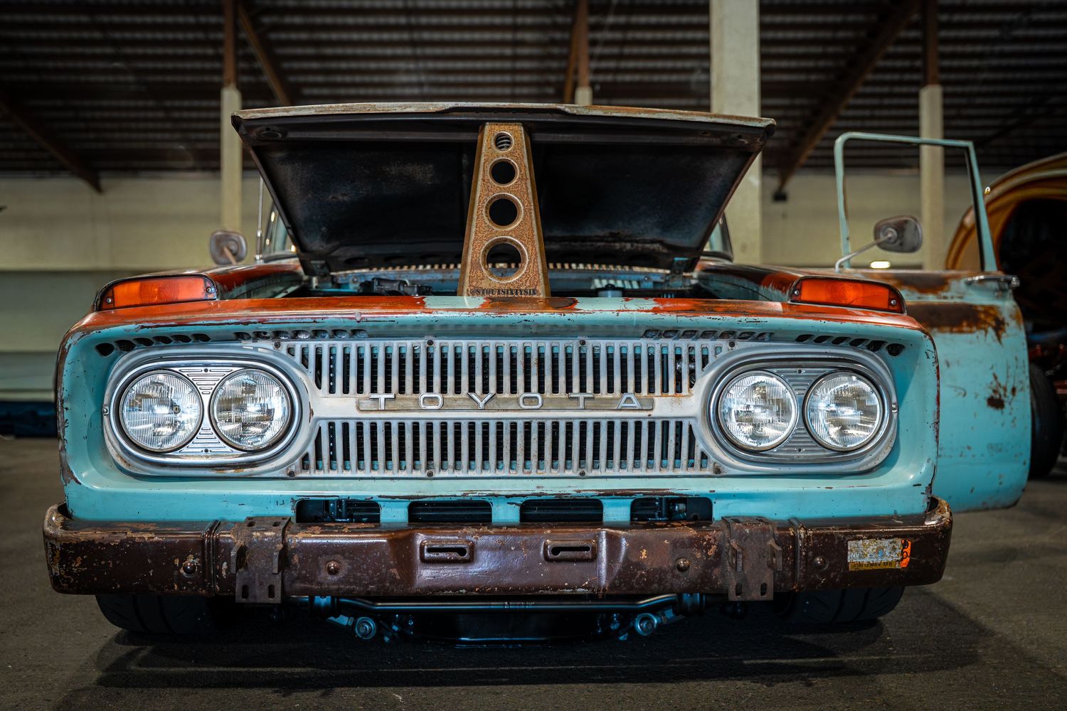 Vintage turquoise truck with rusty patina, open hood, dual headlights, and chrome grille displayed in a garage.