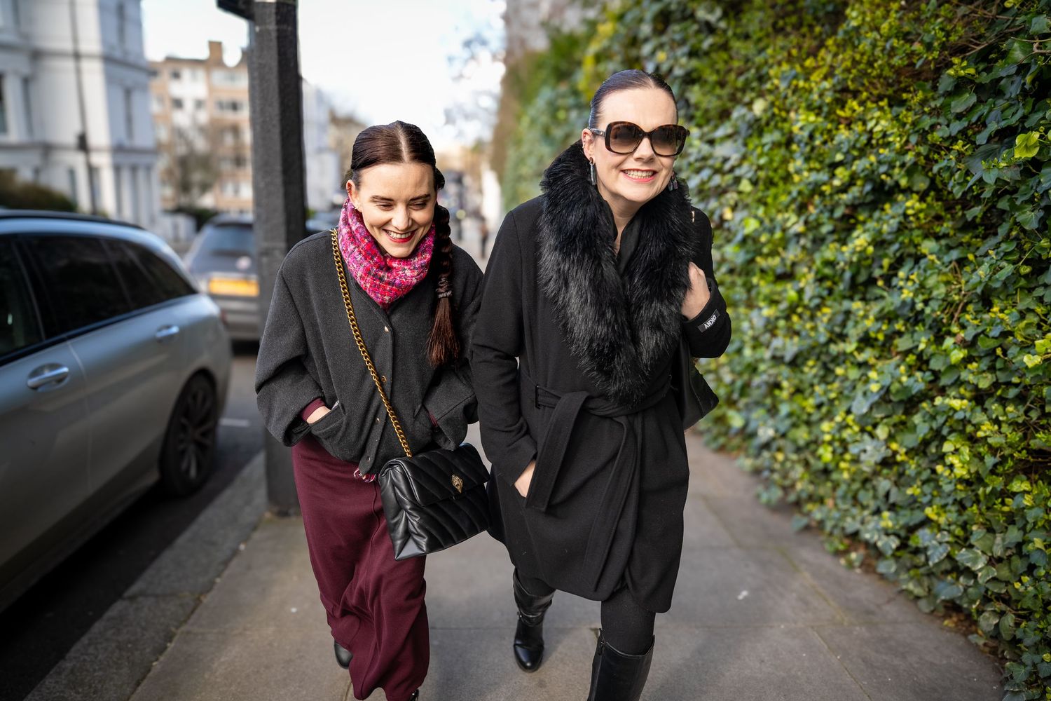 Two people walk together on a city street wearing winter coats and sunglasses on a sunny day.