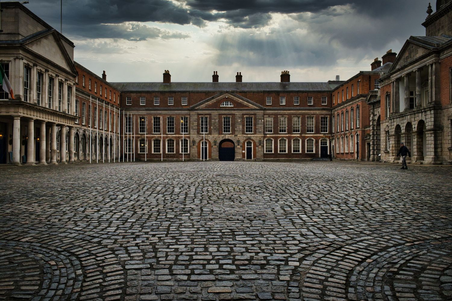Historic cobblestone courtyard of a grand palace complex under dramatic stormy clouds.