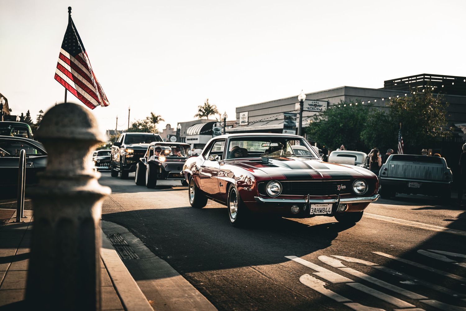 Classic American cars lined up at sunset with US flag visible in background.