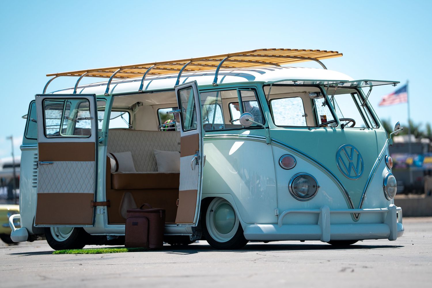 A vintage light blue Volkswagen Type 2 bus with a roof rack, lowered suspension, and open sliding door in a parking lot.