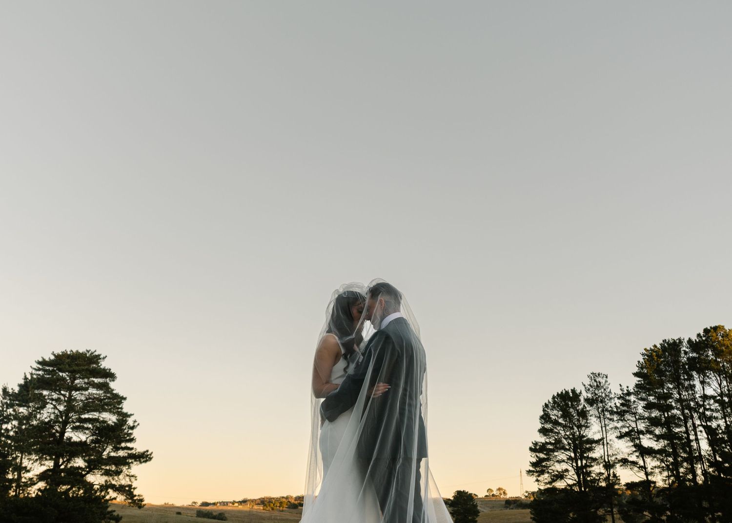 Bride and groom sharing an intimate moment under a veil at sunset with trees in the background.