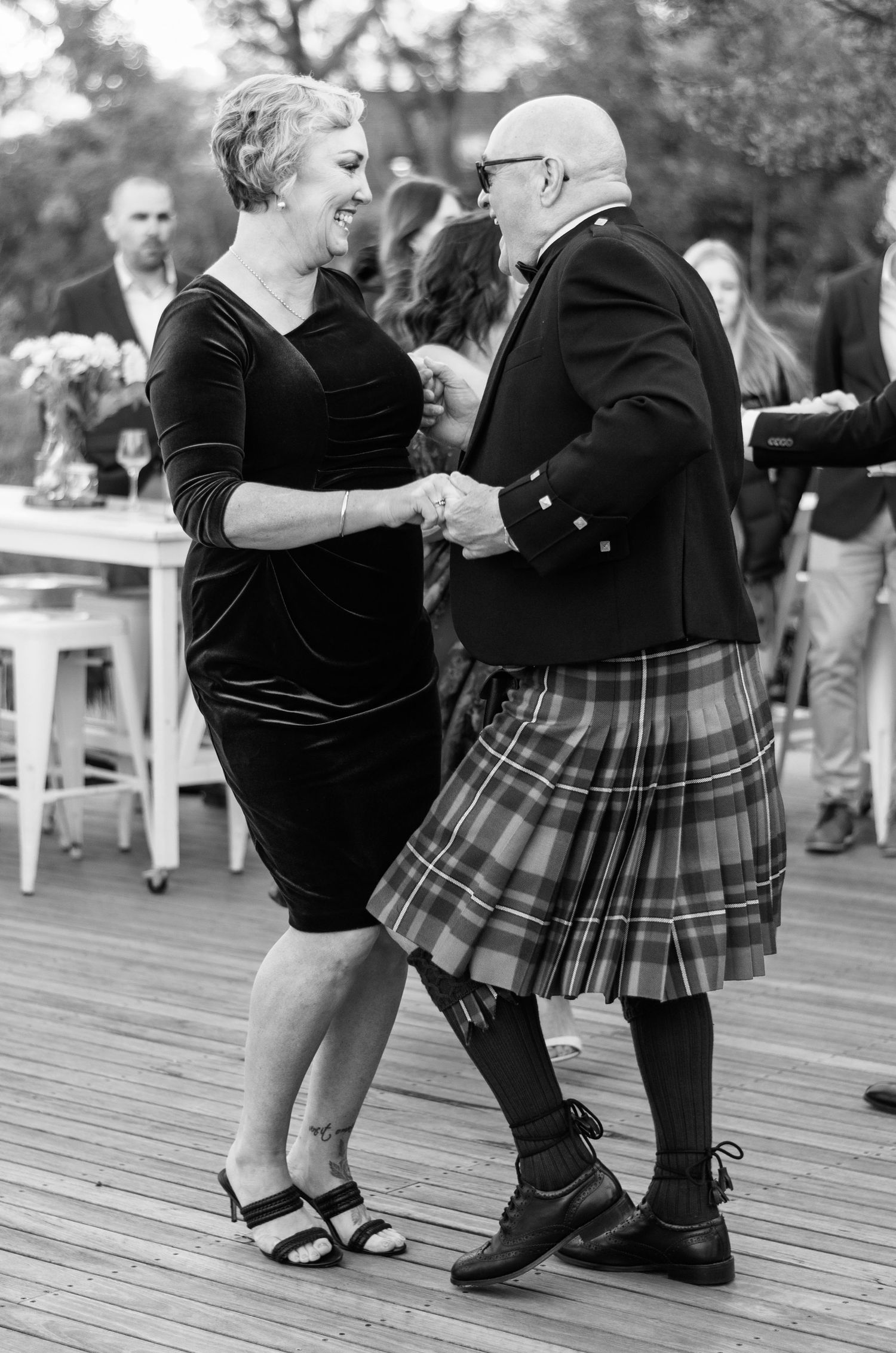 A couple dances joyfully outdoors, the man wearing a traditional Scottish kilt at a wedding reception.