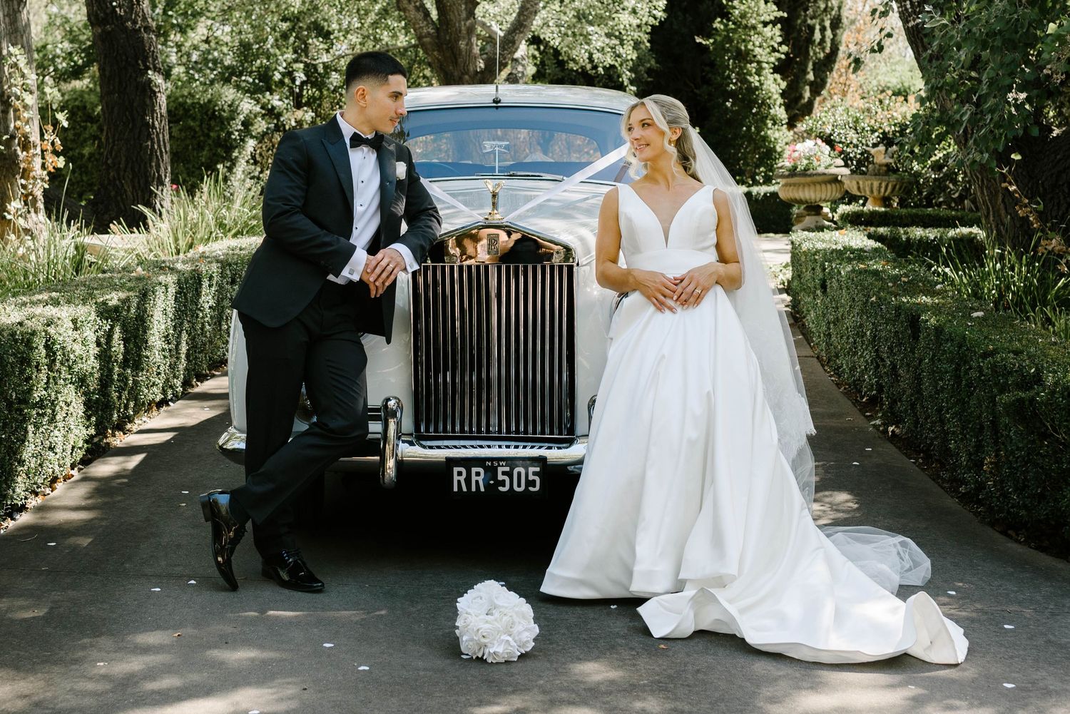 Bride and groom posing beside a vintage white Rolls-Royce RR505 in a lush garden setting on their wedding day.