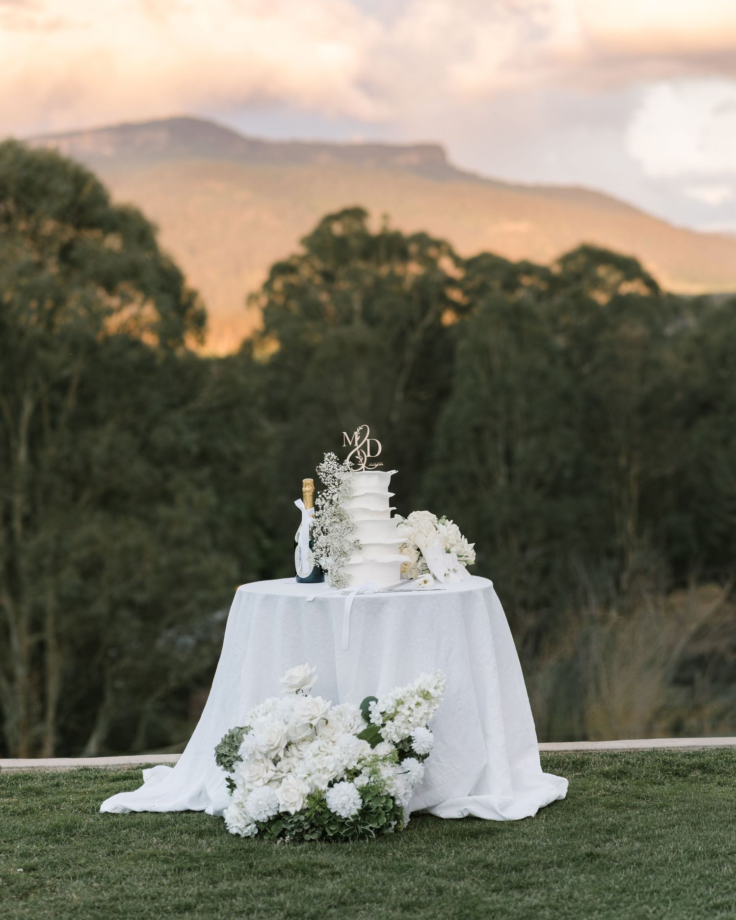 White wedding cake with gold topper on a draped table with white floral arrangements, set against eucalyptus trees and mountains at sunset.