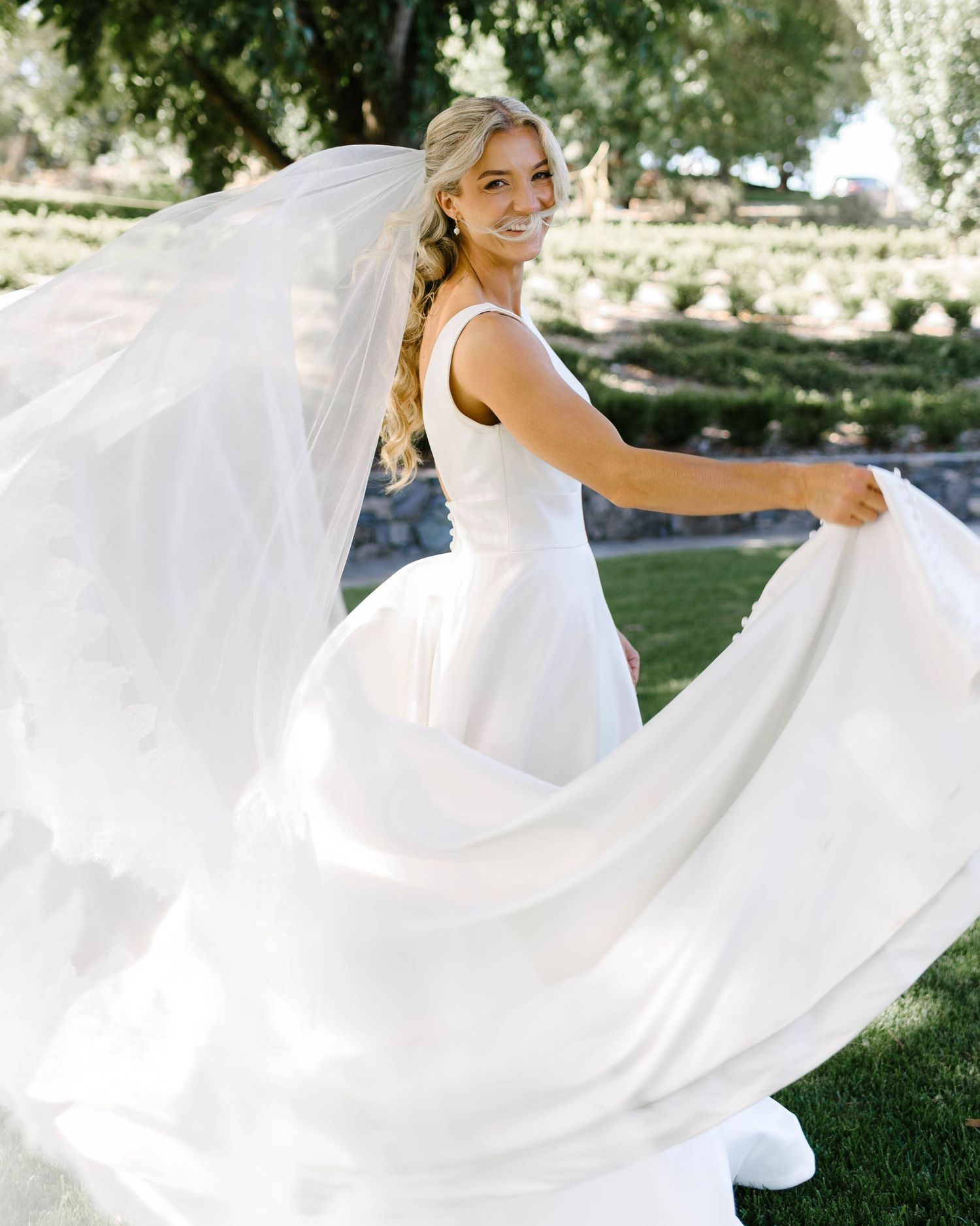 Bride twirling in elegant white wedding gown with flowing veil outdoors at vineyard venue.