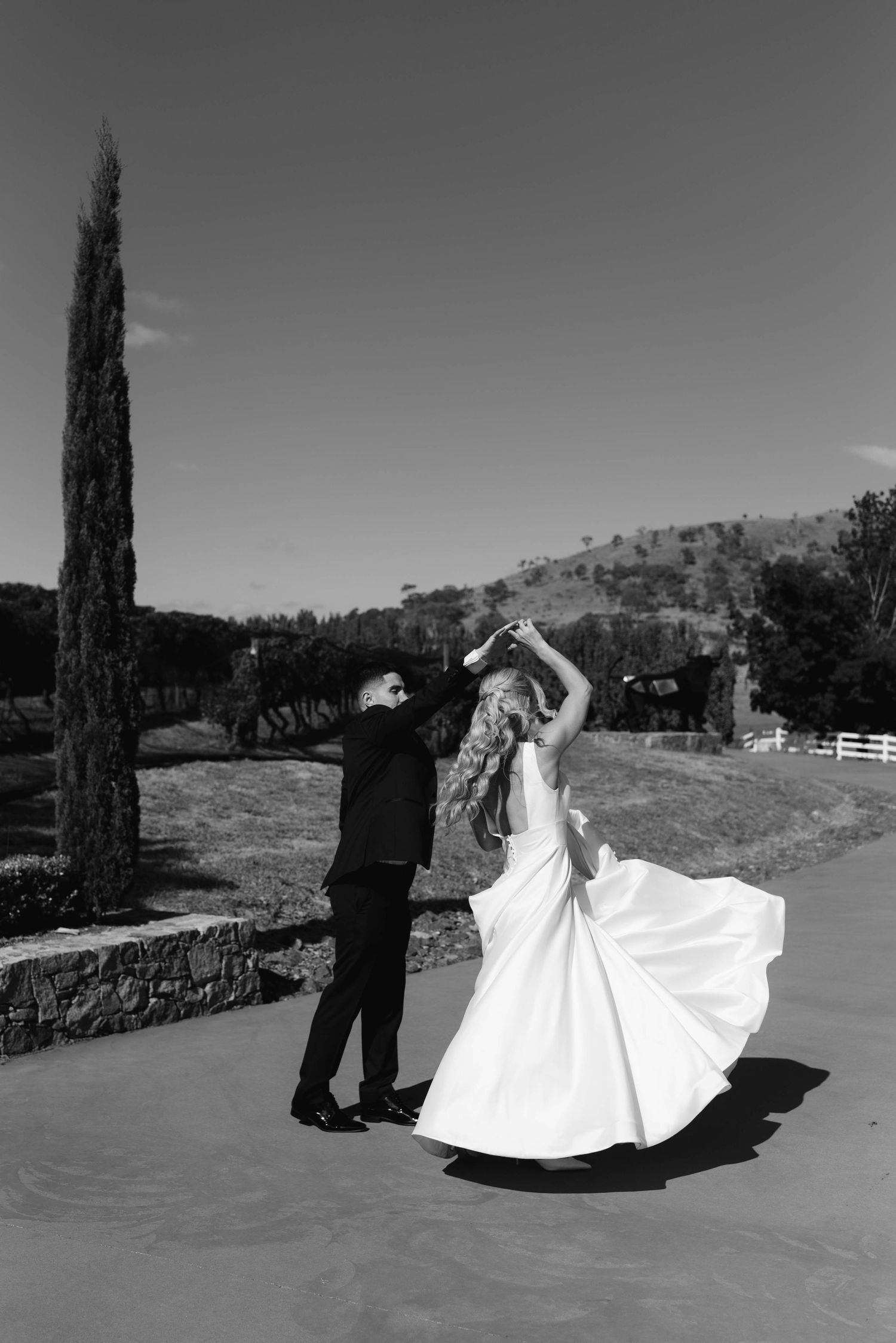 Black and white photo of bride and groom dancing outdoors, with cypress tree and rolling hills in background.