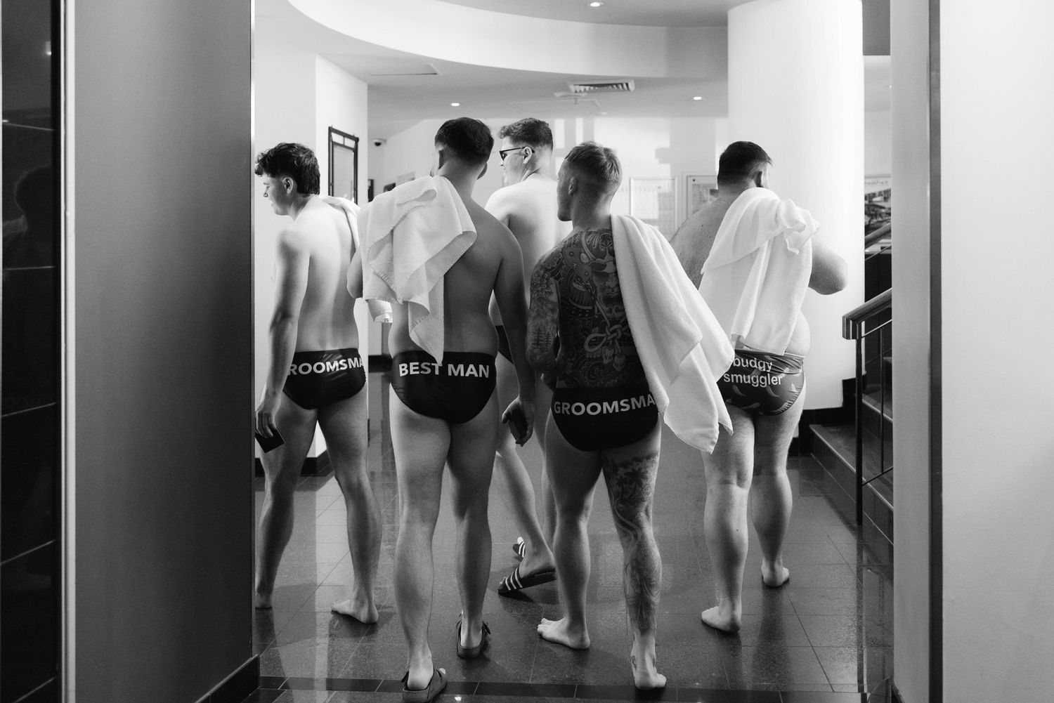 A black and white photograph shows several people walking in a curved hallway with their backs turned to the camera.