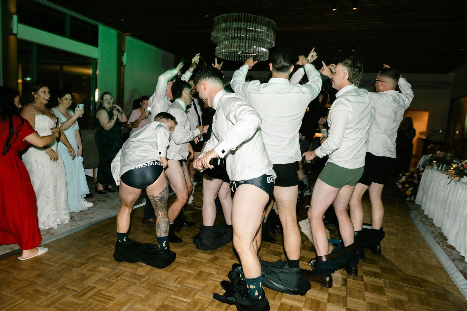 A group of people in matching outfits perform a synchronized dance routine at an indoor event with a chandelier overhead.