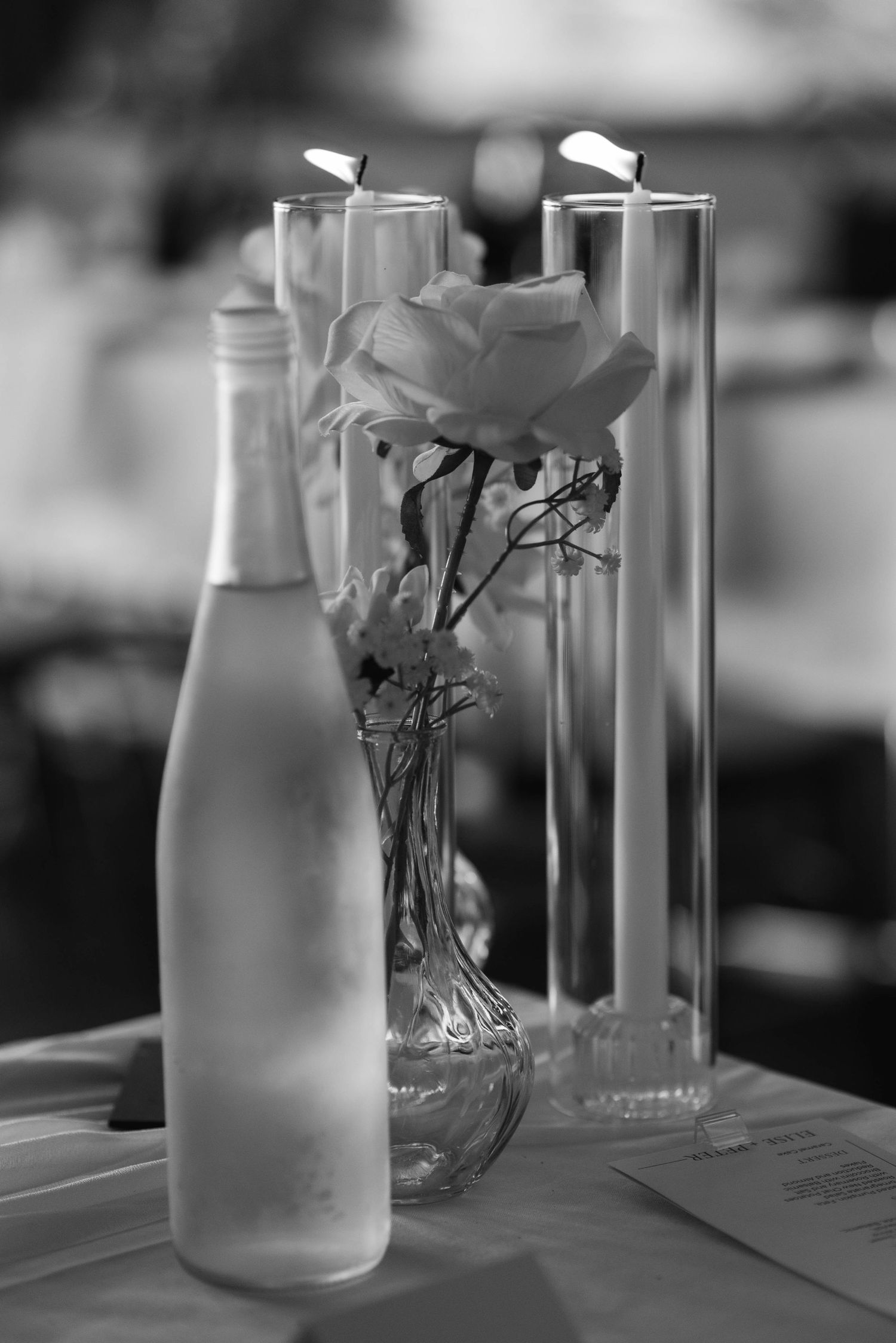 Black and white photo of elegant wedding table centerpiece with roses, candles in glass cylinders, and a decorative bottle.