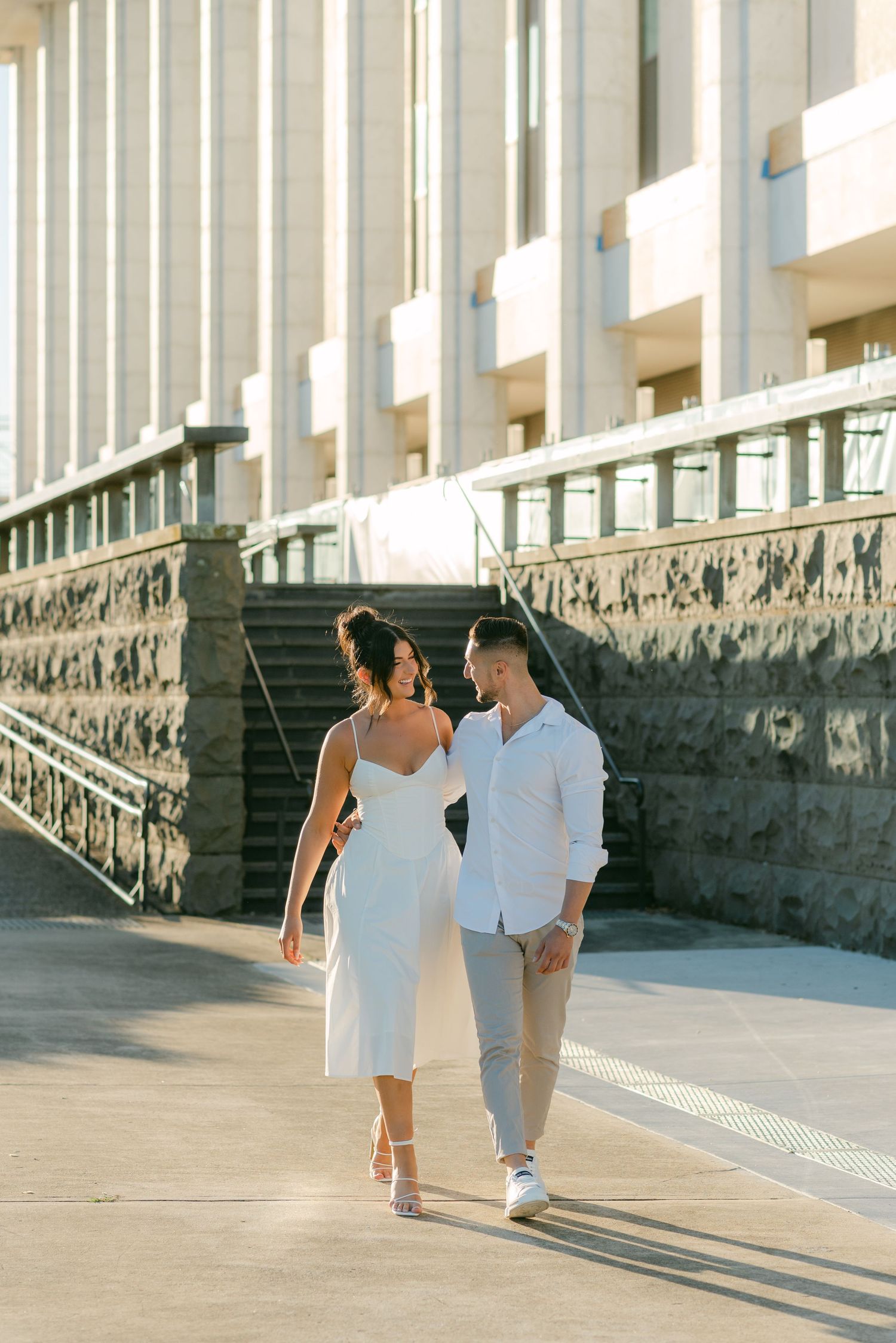 A couple in white outfits walks along a stone-lined path on a sunny day.